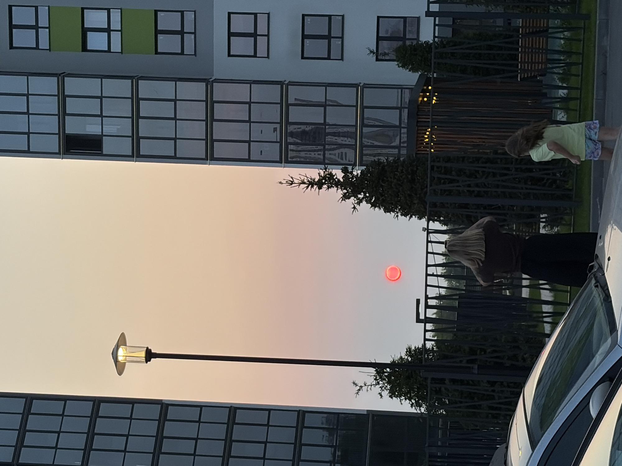 The image captures a street scene in Ozernyy, Russia, during what appears to be sunset or sunrise. Two young girls are present. One, with blonde hair, stands facing away from the camera, looking towards a bright red, circular object in the sky, possibly the sun obscured by atmospheric conditions. She is wearing a brown top and a dark skirt. The other girl, with longer brown hair, is further to the right, facing away from the camera and moving towards the right. She is wearing a light green t-shirt and patterned shorts. They are positioned near a metal fence. Two modern apartment buildings, characterized by their glass-paneled windows and geometric designs, flank the scene. A tall lamppost stands to the left, and evergreen trees are visible in the foreground and behind the fence. A silver car is parked in the lower left corner. The overall environment suggests an urban residential area.