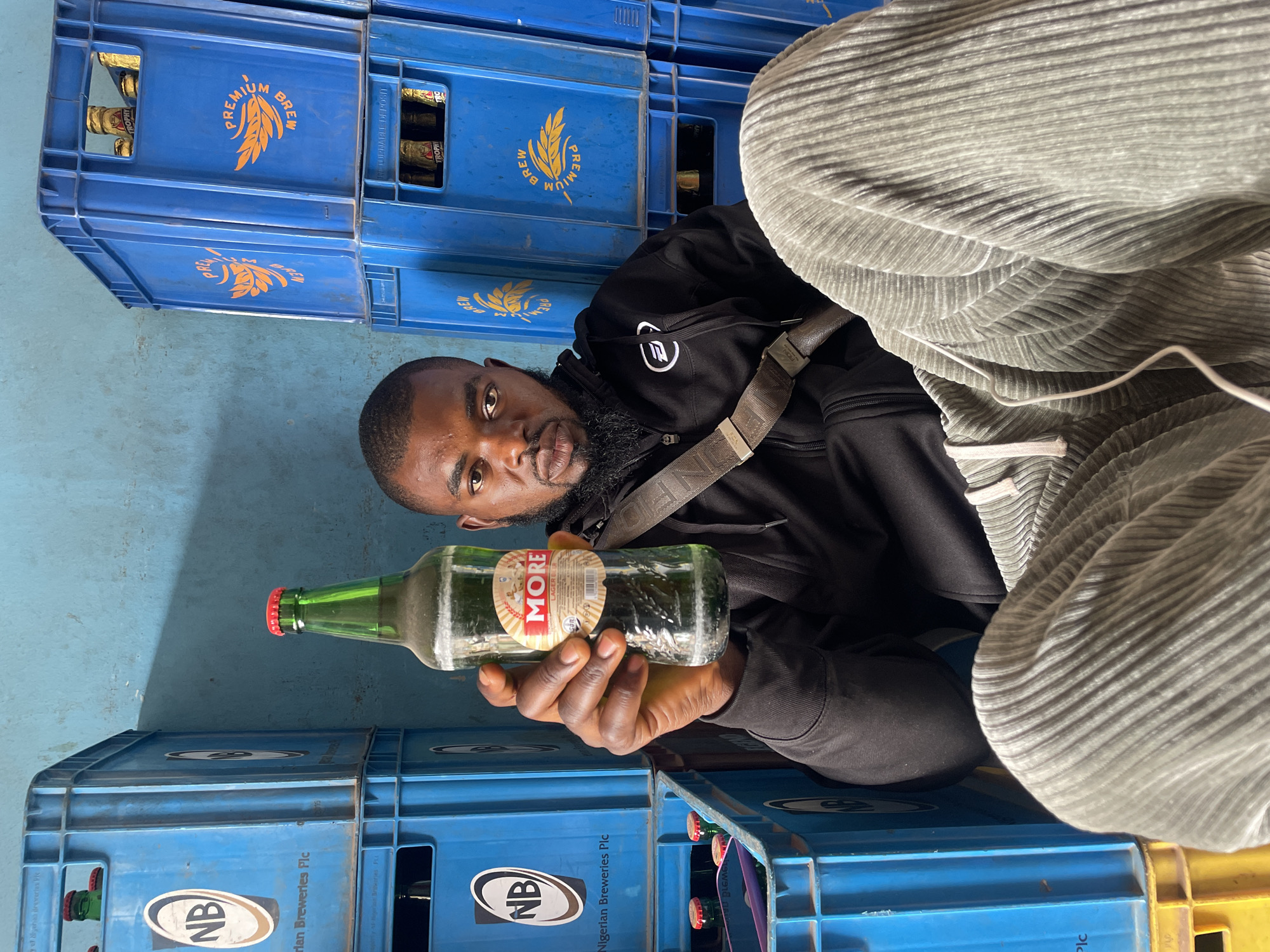 A person is holding a bottle of More Lager Beer in front of a stack of blue plastic crates. The bottle is green and has a red cap. The person has a beard, a black hoodie and is wearing a satchel. The blue crates are labeled "Nigerian Breweries Plc" and have bottles of Premium Brew in them. The location is in Jos, Nigeria.