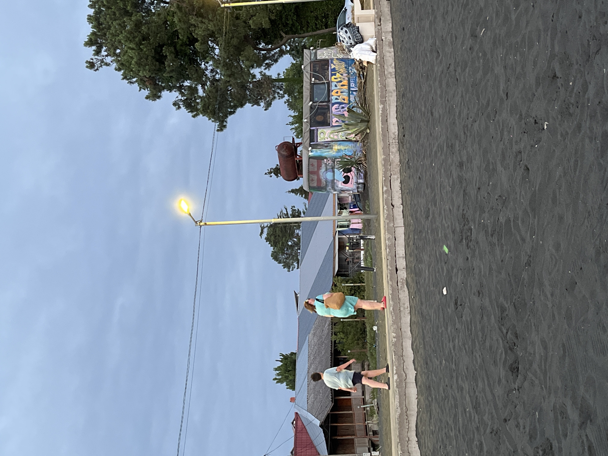 The image shows a view of a black sand beach in Ureki, Georgia, with a promenade in the midground. Two individuals are walking on the promenade, one in a light blue t-shirt and shorts, and the other in a turquoise dress and carrying a large brown bag. To the right of the individuals is a colorful, graffiti-covered bus that appears to be a food or drink establishment, with a large metal tank on its roof. In the background, several buildings with angled roofs and visible wooden structures are present, along with mature trees and a cloudy sky. The overall atmosphere is casual and relaxed, suggestive of a seaside town in the evening or early morning.
