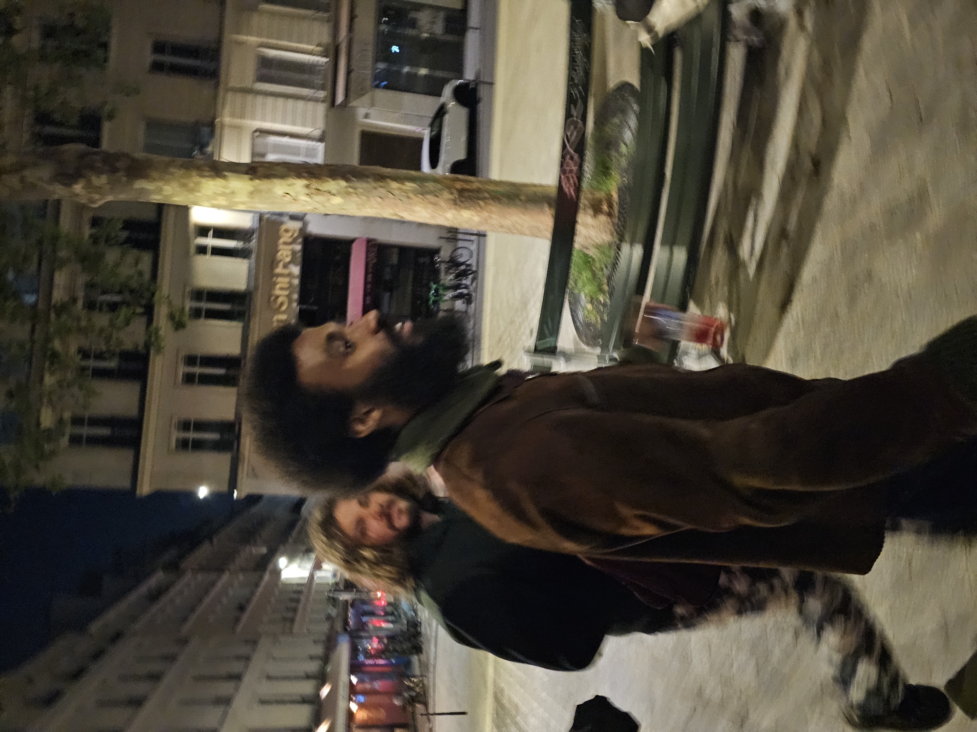 Two people stand on a sidewalk at night in Paris, France. The person in the foreground has dark skin, a full beard, and an afro hairstyle. They wear a brown jacket with a green collar. The person behind them has lighter skin and long blonde hair. They wear a dark jacket over a dark top. The sidewalk is concrete, and there is a metal bench with a green top behind them. A tree trunk is visible in the background, and buildings line the street, including one with a sign that appears to read "shinrang". A car is parked on the street, and there are bicycles nearby. The area is lit by streetlights.