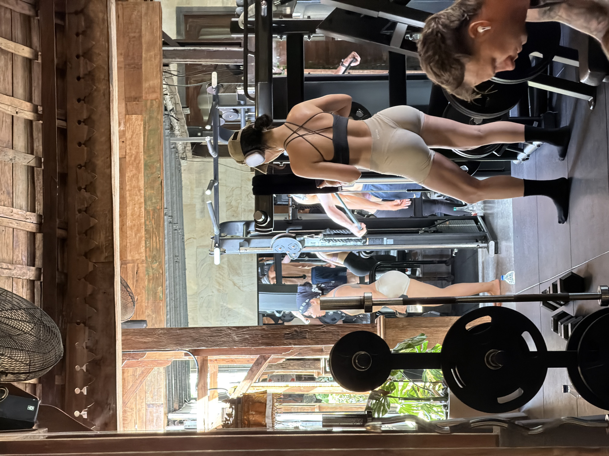 The image depicts a gym setting in North Kuta, Indonesia. Several individuals are exercising on various weight machines. One woman, wearing a black sports bra, light-colored shorts, headphones, and a hat, is pulling on a cable machine. Another person is visible on a separate machine nearby. In the foreground, a barbell with weights rests on a rack. The architectural structure surrounding the gym space features a rustic design with wooden beams and a vintage-style fan. The environment appears to be a blend of modern gym equipment within a traditional Indonesian building.