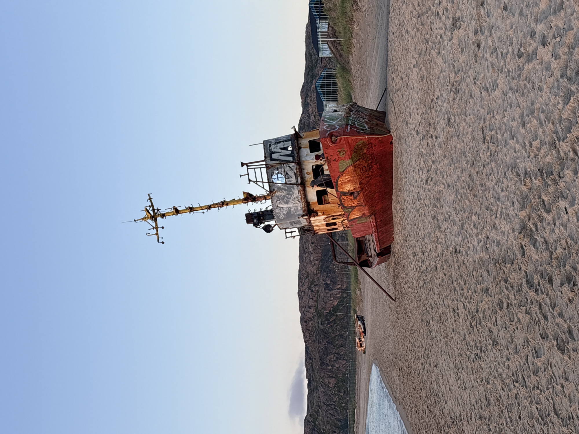 The photograph depicts a rusty, red shipwreck beached on a sandy shore in Teriberka, Russia. The ship, tilted at an angle, exhibits extensive corrosion and graffiti. Its mast and various structures are visible against a clear, pale blue sky. In the background, a rugged, rocky hillside rises, dotted with sparse vegetation. A body of water, likely the Barents Sea, laps at the shore on the left side of the image. Further down the beach, two other smaller, derelict vessels are also visible. Several glass-enclosed structures, possibly greenhouses or storage units, are situated on the slope behind the main shipwreck. The sand is textured with footprints and the overall scene conveys a sense of decay and abandonment.