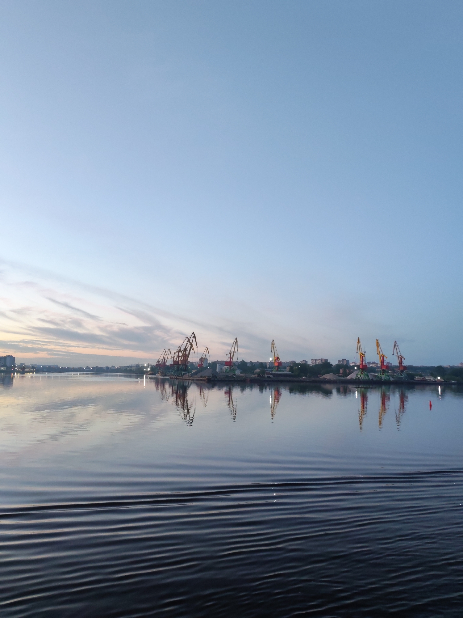The image depicts a tranquil scene on the Sheksna River in Cherepovets, Russia, likely at dusk or dawn. The sky is a gradient of light blue, transitioning to pale orange and pink near the horizon, indicating sunrise or sunset. The river's surface is relatively still, reflecting the sky and the industrial cityscape visible in the background.
The focal point includes a series of large harbor cranes, silhouetted against the skyline, indicating a port or industrial area. These cranes, of varying heights and color schemes (primarily red, yellow, and brown), are reflected in the water, creating a symmetrical, mirror-like effect. Along the shoreline, there is evidence of industrial activity, including piles of materials and some buildings, as well as some trees.
The foreground of the image features ripples on the water's surface, likely caused by a boat or natural water movements. The overall impression is a blend of natural beauty and industrial presence, typical of a river city like Cherepovets. There are no people visible in the frame.