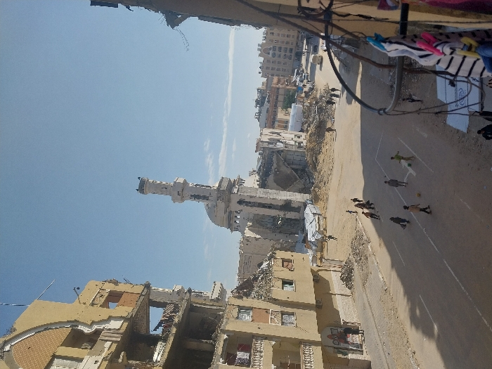 The image depicts a street scene in Khan Yunis, Palestinian Territory, characterized by significant structural damage. In the foreground on the left, a multi-story, light-colored building is severely damaged, with large sections of its facade missing, exposing interior rooms. Its balconies are partially destroyed. In the mid-ground, a prominent mosque features a tall, slender minaret that appears largely intact, while the main mosque structure behind it is heavily damaged or partially collapsed, surrounded by debris. Further down the street and to the right, other buildings are visible, some severely damaged and others appearing relatively intact in the distance.
On the paved street in the mid-ground, approximately 10 to 12 children are engaged in playing soccer with at least two balls. They are casually dressed. Several adults are walking on the sidewalks and further down the street. Piles of rubble and debris are scattered along the sides of the street and around the damaged buildings. A light-colored van is visible on the street in the distance. The sky is clear blue with minimal cloud cover. In the lower right foreground, a clothesline with various items of laundry, including striped garments and colored clothespins, partially obscures the view, suggesting the photograph was taken from an elevated position, such as a balcony.