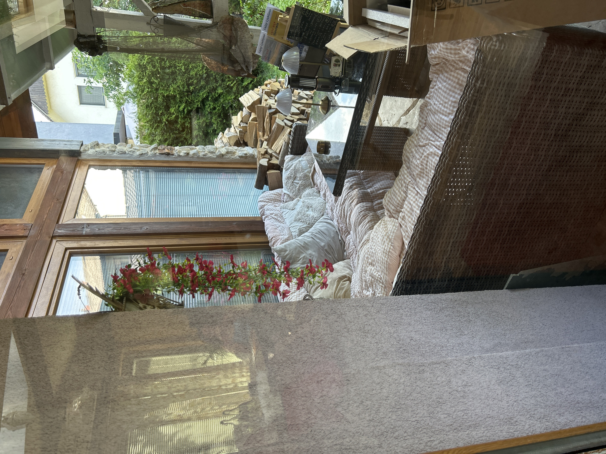 The image depicts an outdoor seating area with a wicker sofa and a glass-topped table. The sofa is covered in light-colored cushions with a checkered pattern. On the table, there are two lamps, a small decorative item, and several papers, including a travel map of Slovenia. Behind the table, a pile of chopped firewood is visible. The area is enclosed by wooden-framed windows and stone walls, and a hanging plant with red flowers drapes from above. In the background, greenery and houses can be seen. The overall impression is of a rustic and comfortable outdoor lounge space.