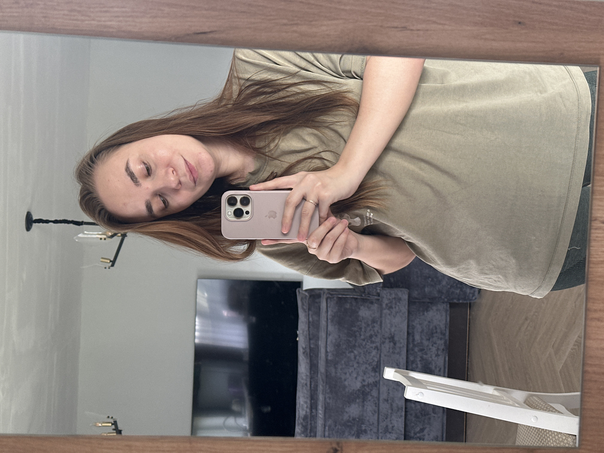 The media file displays a mirror selfie taken by a young woman in what appears to be a residential setting in Kharnavy, Russia. The woman, positioned on the right side of the frame, has long, flowing light brown hair and a light complexion, with a subtle smile. She is wearing a relaxed-fit, light olive-green short-sleeved t-shirt. She holds a light pink iPhone, featuring a visible triple-camera system, in her left hand, which also displays a simple ring on her ring finger. The Apple logo is visible on the back of the phone.
The reflection in the mirror reveals various indoor elements behind her. To the left, a light-colored wall is visible, with a dark, multi-armed chandelier or ceiling light fixture partially reflected. A large, dark flat-screen television is visible in the center-left background, positioned above a dark grey upholstered piece of furniture, likely a sofa or armchair. On the far right, a section of a light-toned wooden floor can be seen, along with part of a white high chair or step stool. The mirror itself is framed by a warm-toned wooden border along its top and bottom edges. The scene captures a casual, indoor moment.