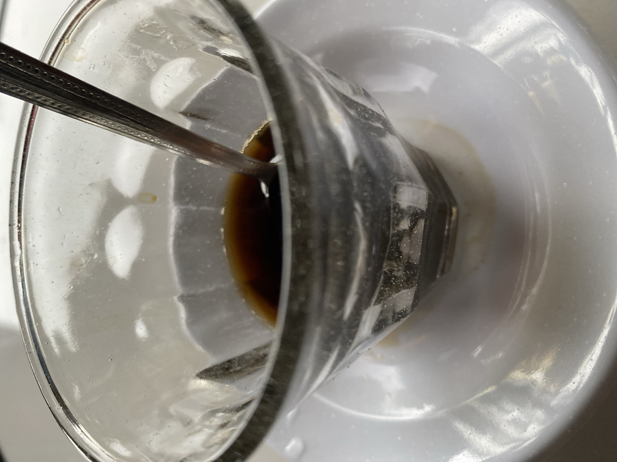 A close-up, overhead shot captures a clear glass bowl, partially filled with dark liquid, resting on a white saucer. A silver spoon is submerged in the liquid, its handle angled upwards. The bowl exhibits subtle ridging, and small air bubbles are visible on its inner surface. The white saucer appears slightly smudged with a light brown residue around the base of the bowl. The background is a soft, out-of-focus white. The scene suggests a moment of consumption, possibly of coffee or tea, in a quiet setting.