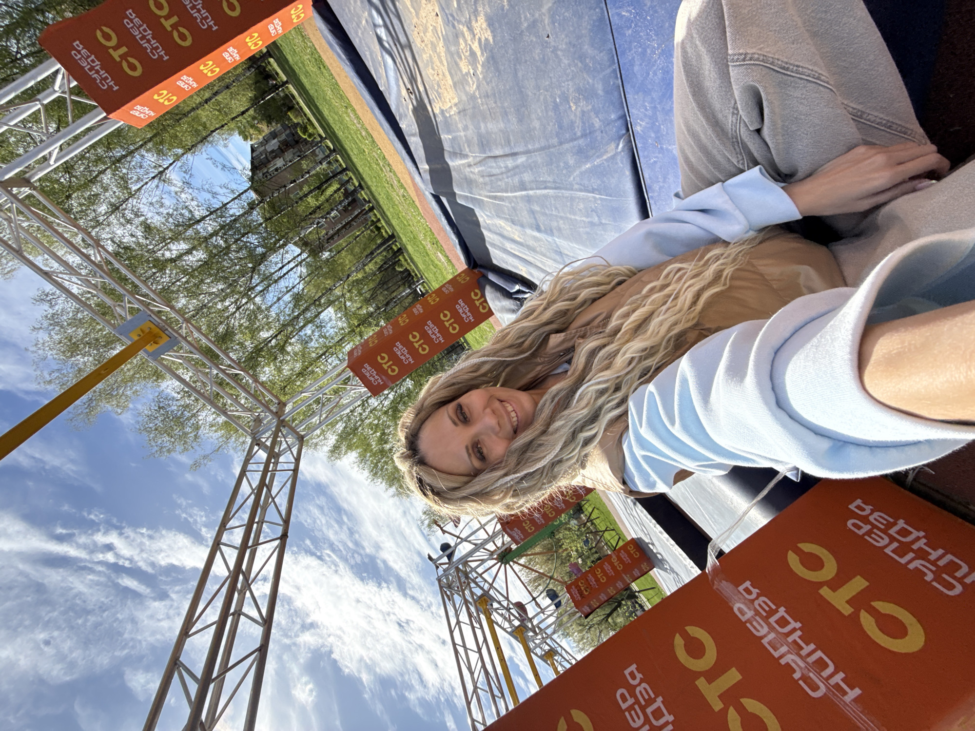 Here is a detailed description of the image:
**Scene:** The image is a selfie taken from a low angle, capturing a woman sitting on or in what appears to be a part of an obstacle course or amusement park attraction.
**People:** A woman with long blonde hair, wearing a light blue top and light-colored jeans or pants. She is smiling towards the camera.
**Objects:** There are several orange signs with the Russian text "CTC СУПЕР НИНДЗЯ" (CTC SUPER NINJA) displayed on them. These signs appear to be part of the obstacle course or event setup. There are metal structures, possibly part of a frame or support for the attraction. The background includes greenery, possibly trees and grass, and a blue and cloudy sky.
**Environment:** The setting is outdoors, possibly in a park or recreational area. The presence of the obstacle course and signage suggests a competition, amusement park, or event setup.
**Location:** The signs indicate the event is related to "CTC СУПЕР НИНДЗЯ", and the OCR indicates that the location is likely in Шебунино, Russia.