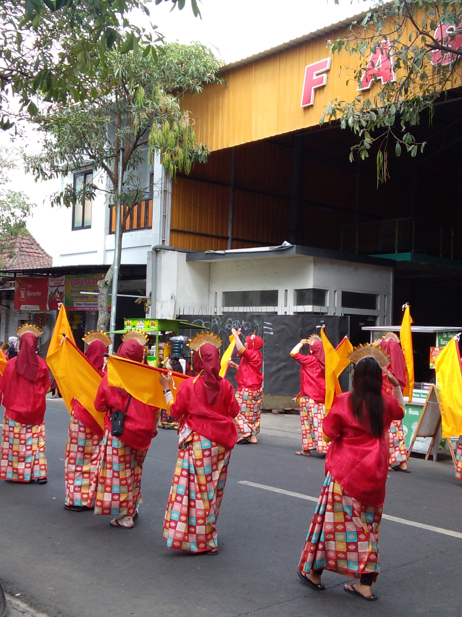 This is a photograph of a group of people participating in a parade on a street in Trenggalek, Indonesia. The people are dressed in traditional red outfits with elaborate patterned skirts and gold headpieces. They are holding yellow flags and appear to be dancing or performing. The parade is taking place in front of buildings, one of which has a large yellow facade with red lettering. The street is paved and there are trees and other foliage visible. The overall atmosphere suggests a festive cultural event.