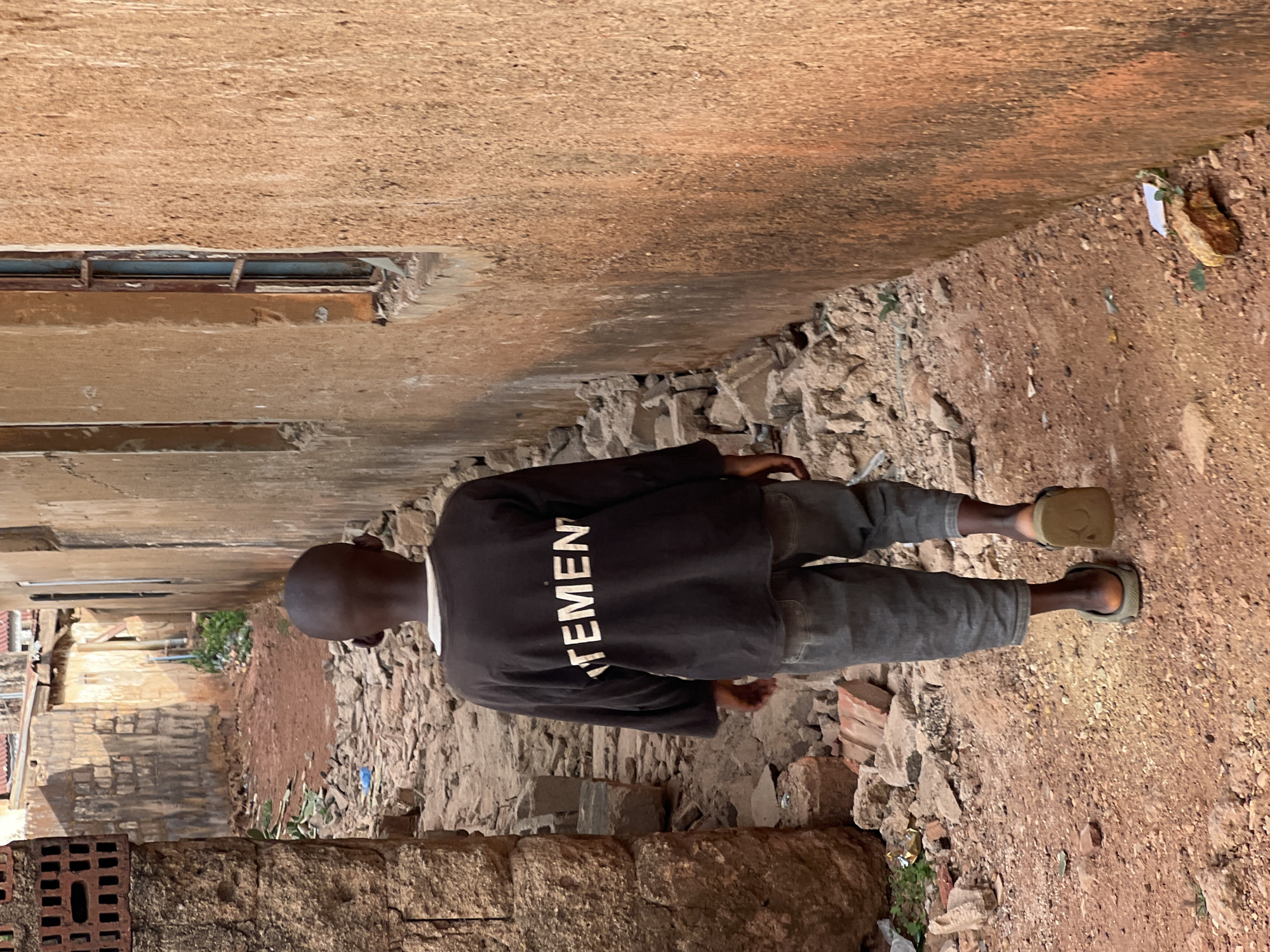A young, slender person stands outdoors in Gura Top, Nigeria. He faces away from the viewer in a narrow, rubble-strewn alley between buildings. The person wears a dark, long-sleeved shirt with a white word vertically printed on the back, dark pants, and sandals. The ground is dirt and loose stones, with some brick debris. The buildings are of brick and mortar, with various surface imperfections and some small windows. The environment is uneven and possibly unpaved.