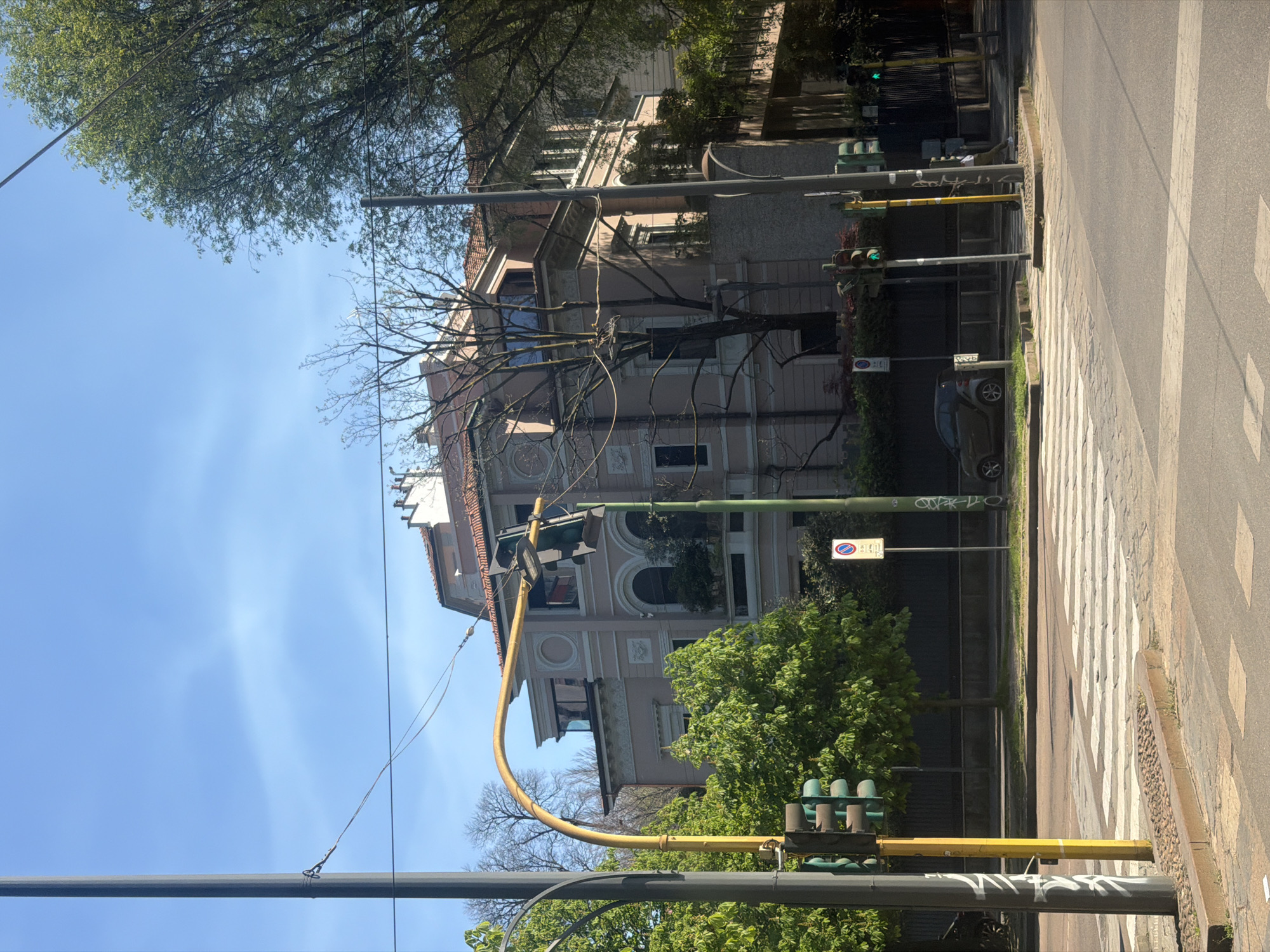 The image depicts a street scene in Milan, Italy. A three-story building with an ornate facade is the main architectural feature. It appears to be a residential structure, possibly an apartment building. A tree with bare branches partially obscures the building's facade.
On the right side of the image is a pedestrian crosswalk and a street. A traffic light stands at the intersection, displaying a green light. A car is parked along the side of the street. A "no parking" sign is visible. Above, the sky is blue, with a few wisps of clouds. The image is taken from street level.