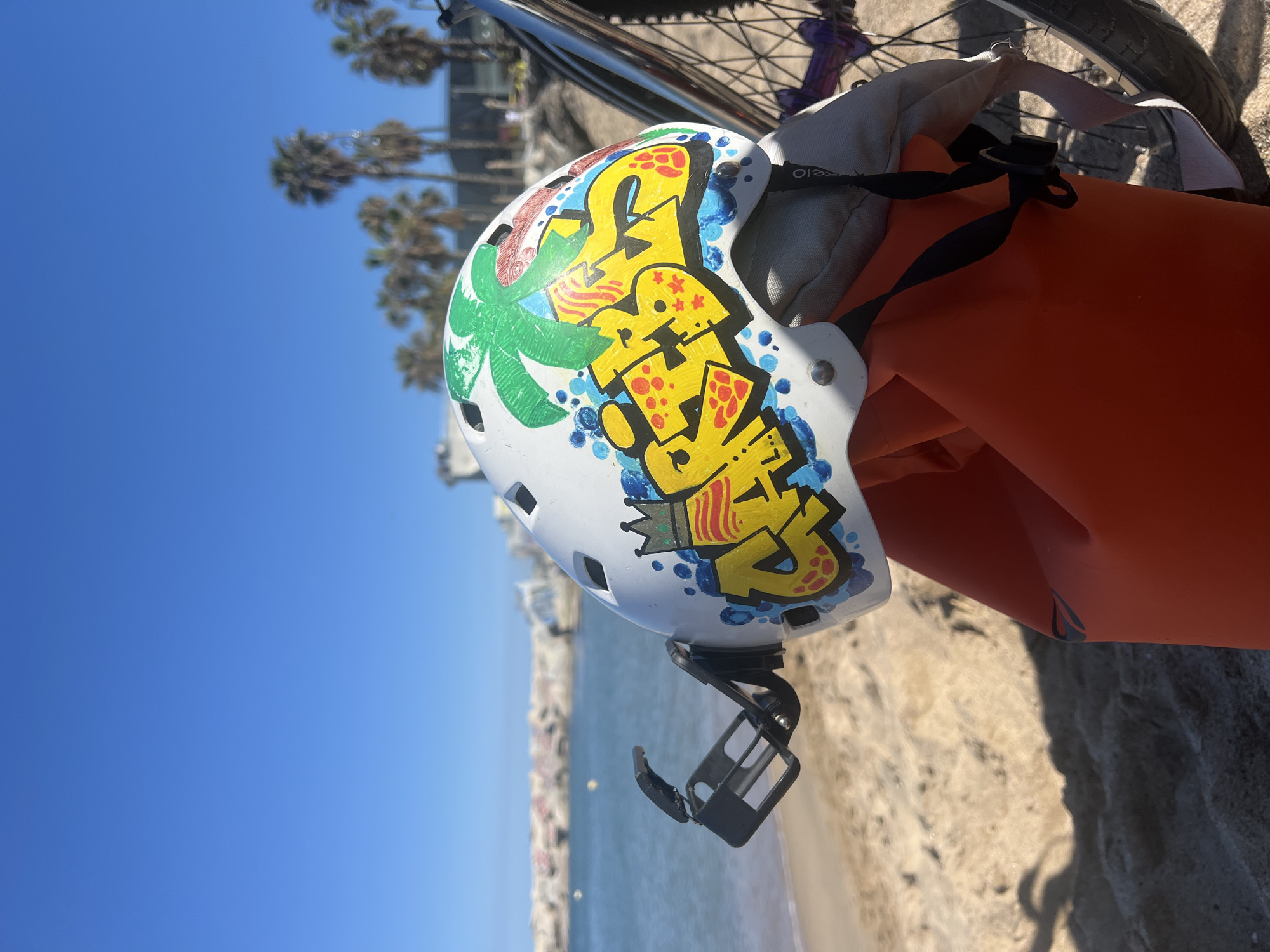 A bicycle helmet rests on an orange bag, positioned on a beach with palm trees and the sea visible in the background under a clear blue sky. The helmet is white and adorned with colorful graffiti, including a palm tree and the word "SUMER". A black camera mount is attached to the helmet. The setting appears to be a beach, likely in a coastal area, possibly in Barcelona, Spain.