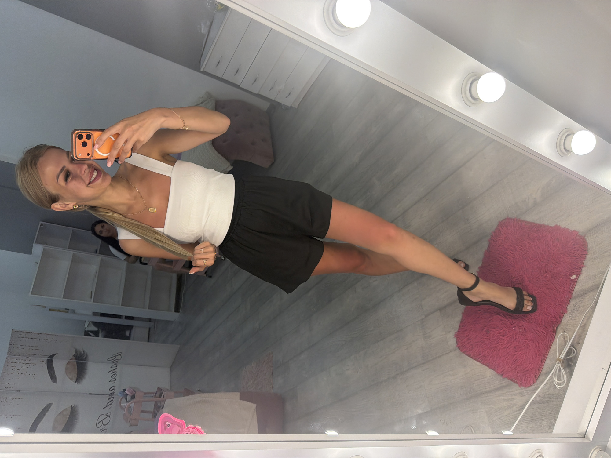This media file shows a young, blonde woman taking a mirror selfie in what appears to be a beauty salon or studio. She is smiling brightly at the camera, holding an orange-cased smartphone. She is wearing a white sleeveless top and black shorts. Her left leg is extended, and she is wearing dark, strappy sandals on her right foot, which rests on a small, fluffy pink mat. The background of the mirror reflection reveals a room with shelves, a chair, and a wall adorned with images of eyelashes, suggesting a focus on beauty services like lash extensions. The setting is well-lit, with a series of light bulbs visible around the mirror, indicating an indoor environment. The overall mood is cheerful and confident, conveyed through the woman's smile and pose. The image is shot in portrait orientation.