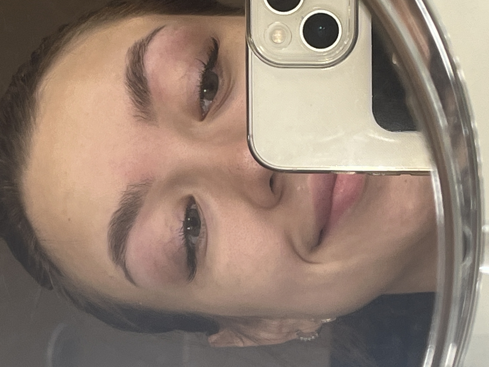 The image is a close-up, self-taken photograph of a young woman's face, reflected in a mirror. The woman has dark eyebrows, dark eyelashes, and appears to be smiling. She is wearing subtle makeup, with pinkish hues on her eyelids and cheeks. A portion of a smartphone, held by the woman, is visible in the lower right of the frame, with its camera lenses pointed towards the mirror. The background is dark and indistinct. The photograph is taken indoors. No specific location in Chaikovsky, Russia, is identifiable from the visual information provided.