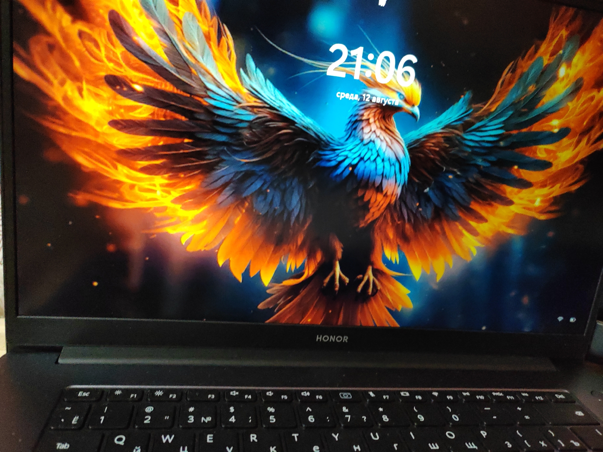 A laptop displays a vibrant wallpaper of a phoenix with fiery orange wings spread wide against a dark blue, star-filled background. The phoenix has turquoise and black feathers on its body and wings, and a yellow and orange head. The time "21:06" is displayed in white text on the screen, below which is the date "среда, 12 августа" (Wednesday, August 12) in smaller white text. The "HONOR" logo is visible below the screen on the laptop's bezel. The keyboard of the laptop is in focus in the foreground. The scene is indoors, with no specific city or location details discernible.