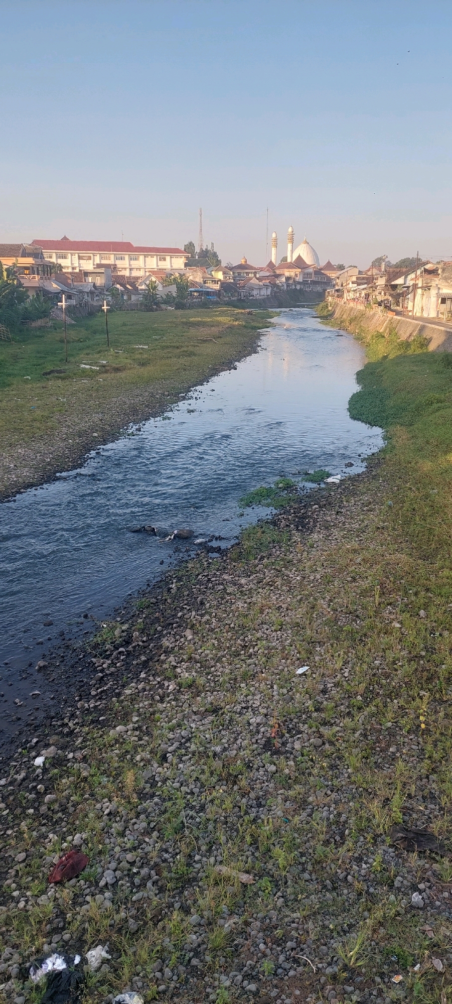 The image shows a view of the city of Lumajang, Indonesia, with a river flowing through it. The river has rocky banks covered in sparse green grass. On the left bank, there is a grassy field with two wooden posts. On the right bank, the terrain is steeper, with more grass and a concrete pathway above. In the background, buildings line the horizon, including what appears to be a mosque with a large white dome and two minarets. The sky is clear and blue, suggesting it is daytime. The scene appears to be a relatively calm, urban river setting with some evidence of human activity and settlement.