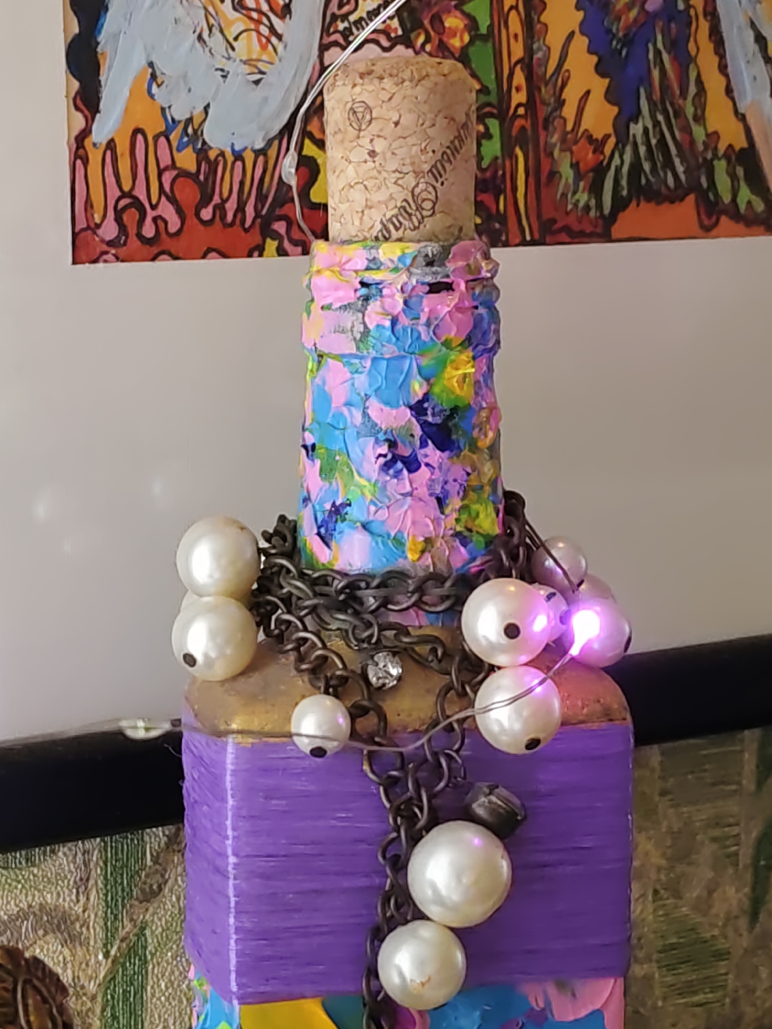 The image shows a close-up of a decorated bottle with a cork stopper. The bottle is adorned with colorful paint, a chain, and pearl-like beads. Some of the beads are lit with pink lights. The background includes a portion of a colorful painting. The specific location in Rostov-on-Don, Russia, cannot be determined from the provided image.