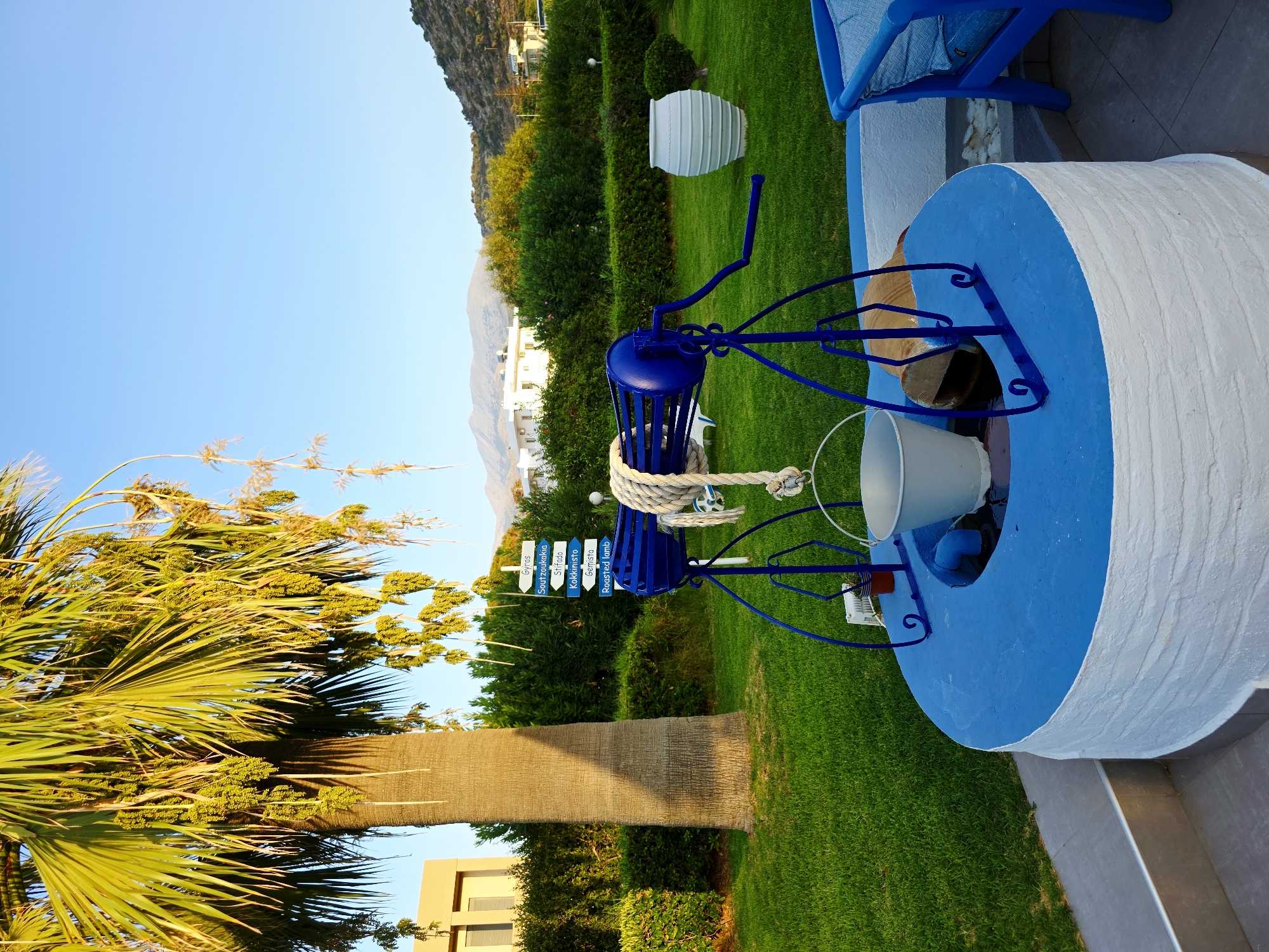 An outdoor scene in Malia Municipal Unit, Greece, features a decorative well with a blue metal mechanism for drawing water. A white bucket hangs from the mechanism. The well is surrounded by a low, white, textured wall with a bright blue rim. Behind the well, a lush green lawn stretches towards a backdrop of dense green bushes and trees. In the distance, white buildings and hills are visible under a clear blue sky. To the left of the well, a tall palm tree with hanging fruit shades a portion of the lawn. Several directional signs are mounted on a pole near the palm tree, indicating "Gyros," "Soutzoukakia," "Stifado," "Kokkinisto," "Gemista," and "Roasted lamb." A large white amphora-shaped vase stands to the right of the well, and a blue chair with a cushion is also present. The overall impression is one of a well-maintained garden or courtyard.
