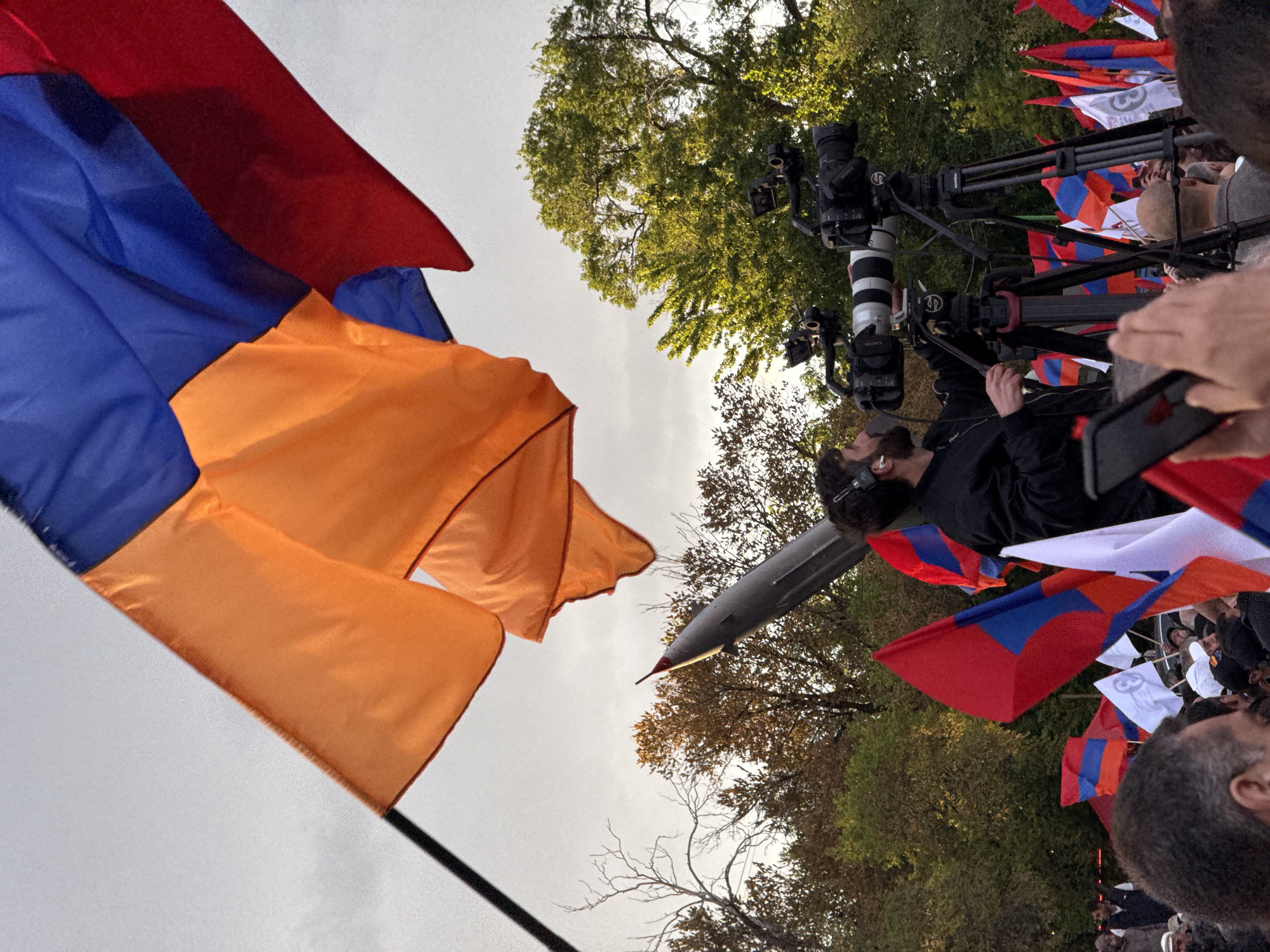 The image depicts a protest or demonstration, likely in Yerevan, Armenia, given the prevalence of Armenian flags. The scene is outdoors, possibly in a park or public space, as indicated by the presence of trees. The sky is overcast.
Several people are visible, including a cameraman with a professional camera and tripod, suggesting media coverage. The cameraman is wearing headphones and is focused on capturing footage. Other individuals are partially visible, indicating a crowd.
Numerous Armenian flags are being waved or held up, creating a sea of red, blue, and orange. There are some white flags present. In the background, there is what appears to be a fighter jet or missile mounted on a pole.