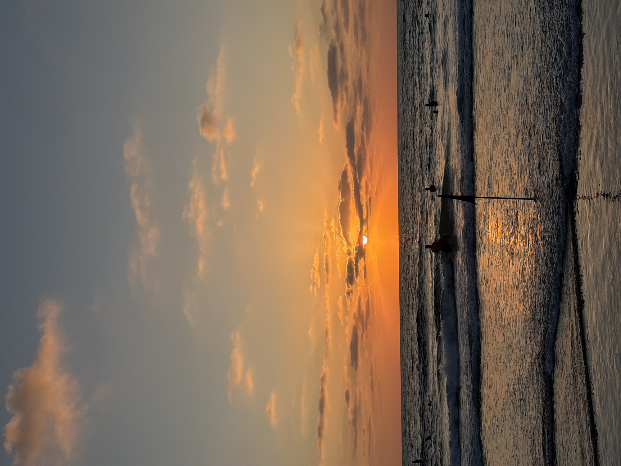 A vertical, full shot captures a vibrant sunset over the Mediterranean Sea at Tel Aviv's Gordon Beach. The sky transitions from a soft blue at the top to a fiery orange near the horizon, with scattered, wispy clouds catching the light. The sun, partially obscured by clouds, casts a brilliant golden glow across the water, illuminating the gentle waves rolling onto the shore.
In the shallow water, a lone surfer, silhouetted against the sunset, is paddling on a surfboard. Further out, several other figures are visible in the sea, some appearing to be swimming. A small black flag on a pole stands in the surf near the foreground, adding a subtle human element to the natural spectacle. The wet sand reflects the warm hues of the sky, creating a sense of depth and tranquility. The overall atmosphere is one of peaceful serenity as the day draws to a close.