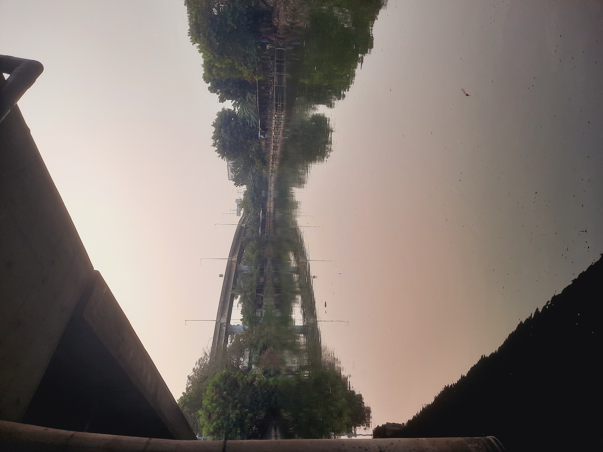 This image depicts a view of the Sunter River in North Jakarta, Indonesia, as seen from beneath an elevated highway overpass. The river's surface is calm, reflecting the overcast sky and the surrounding greenery. On the far bank, a line of stilted wooden structures and trees are visible, suggesting a densely vegetated area with some form of human habitation or activity. A large concrete bridge, part of the elevated highway, stretches across the scene in the mid-ground, its structure casting a shadow onto the water and vegetation. The overall atmosphere is muted, with soft, diffused light, typical of an early morning or late afternoon in a tropical climate. There are no discernible people or specific actions taking place in the scene, which primarily focuses on the natural and infrastructural elements of the urban landscape.