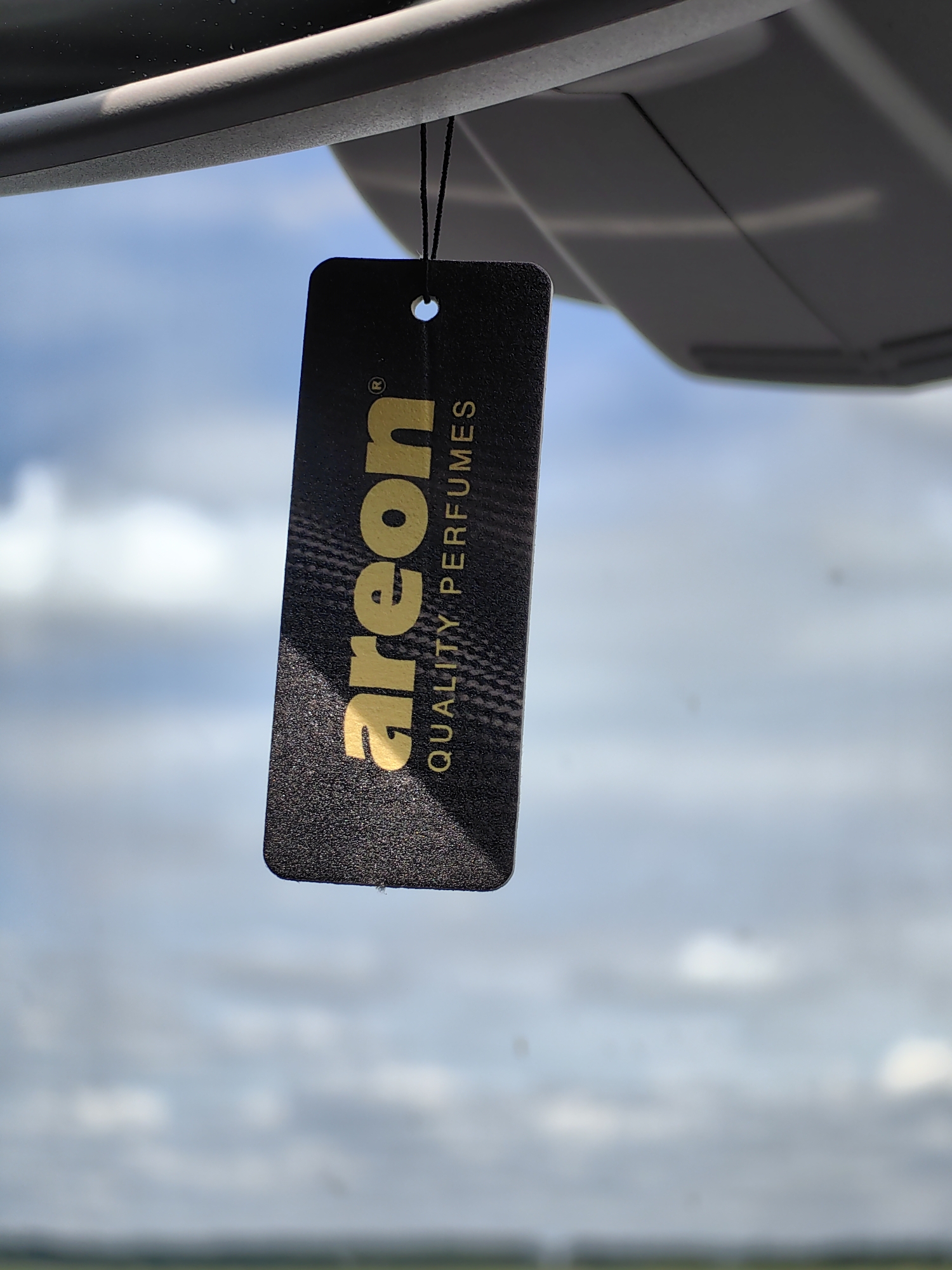 The image, reportedly captured in Дабрынёўскі сельскі Савет, Belarus, displays a black, rectangular car air freshener tag hanging against a blurred outdoor background. The tag is oriented vertically, featuring the brand name "areon" in prominent gold, stylized script, with a registered trademark symbol (®) positioned above the final 'n'. Below this, the phrase "QUALITY PERFUMES" is printed vertically in smaller, gold capital letters. The surface of the tag exhibits a fine, granular texture. A thin black string passes through a circular hole near the top of the tag, leading upwards to an unseen attachment point on a gray, molded plastic structure partially visible in the upper frame. This structure, likely part of a vehicle's interior (such as a sun visor or rearview mirror mount), displays subtle contours and seams. The background consists of an out-of-focus blue sky with scattered white clouds, and a faint green line indicating a distant horizon at the bottom. The scene is illuminated by natural daylight. No individuals or other distinct subjects are present.