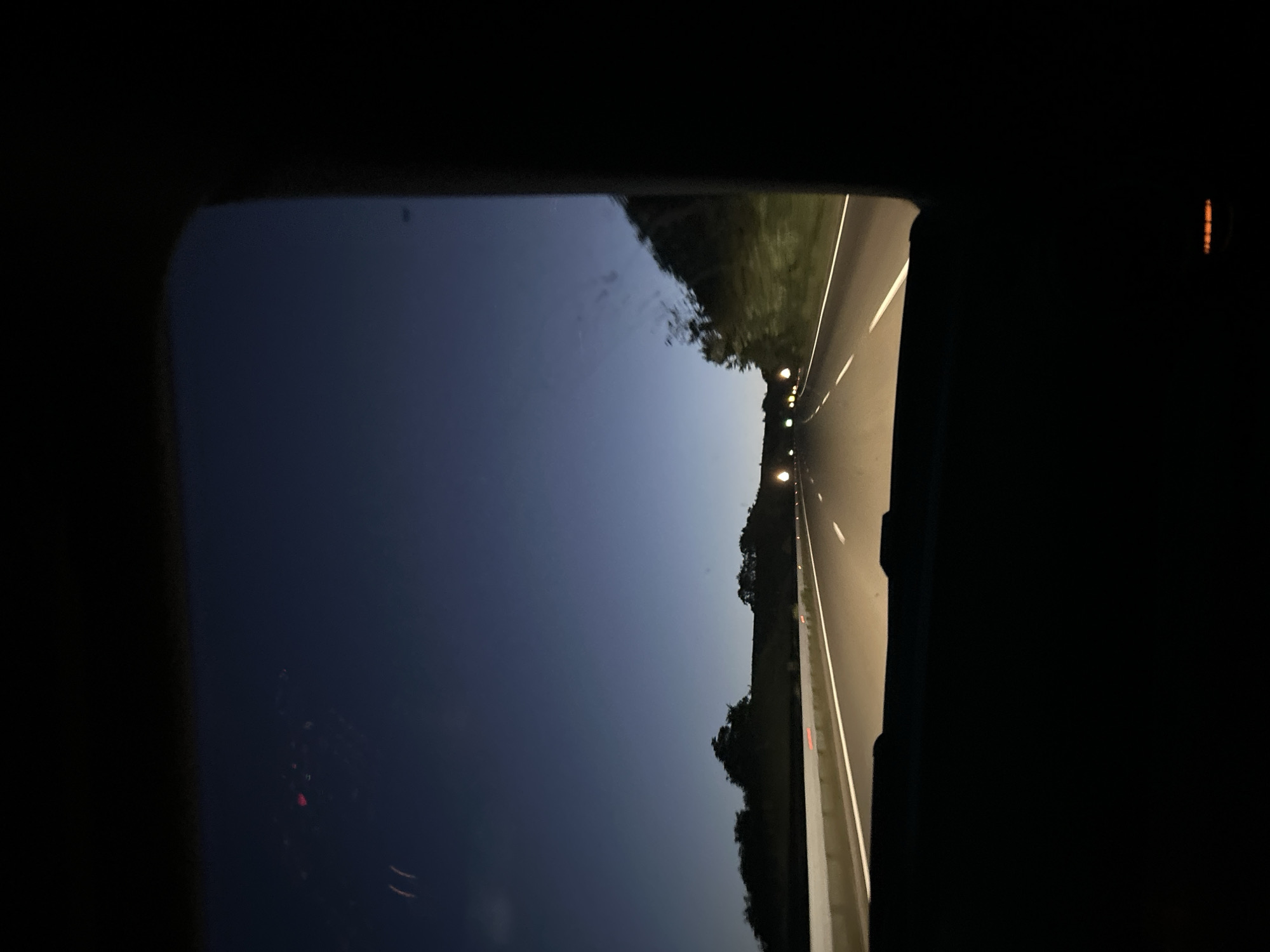 A low-angle photograph taken from inside a vehicle traveling on a road at night. The perspective is through the vehicle's window, with the frame of the window visible on the left and right edges of the image. The road is illuminated by overhead lights and the headlights of the vehicle, with green vegetation visible on both sides of the road. The sky above is dark blue. The specific location within the city of Blatino, Bulgaria cannot be determined from the image.