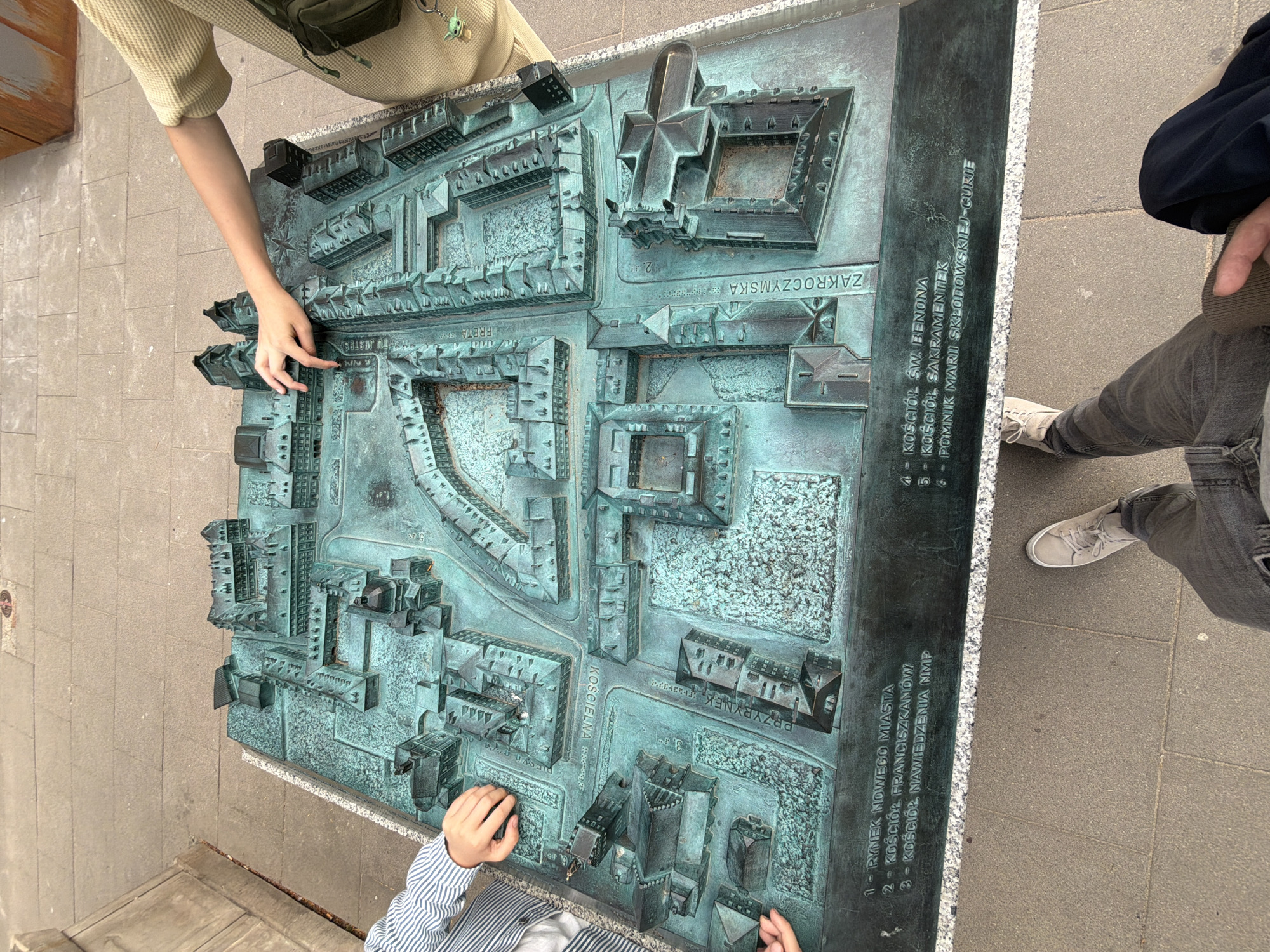 This media file shows a top-down, slightly angled view of a detailed bronze architectural model of a city district, likely an old town, set on a sidewalk. Two individuals are interacting with the model, their arms and hands visible. One person, wearing a light-colored waffle-knit top, points to a section of the model with their right hand. Another person, wearing a blue and white striped shirt, touches a different part of the model with both hands. The model itself depicts numerous buildings of varying architectural styles, streets, and courtyards, all rendered in intricate detail. The bronze has a patina, suggesting it is outdoors and has been exposed to the elements.
The setting appears to be a public space with a paved sidewalk. The lighting suggests it is daytime, possibly overcast as there are no strong shadows.
Visible text on the side of the model's base is inscribed vertically. Portions of this text are legible and appear to be in Polish, listing numbered points. The visible text includes: "RYNEK NOWEGO MIASTA", "KOŚCIÓŁ FRANCISZKANÓW", "KOŚCIÓŁ NAWIEDZENIA NMP", "KOŚCIÓŁ ŚW. BENONA", "KOŚCIÓŁ ŚW. SAKRAMENTEK", and "POMNIK MARII SKŁODOWSKIEJ-CURIE", along with what appears to be "ZAKROCZYMSKA". These indicate specific landmarks and streets within the depicted area. The interaction with the model suggests an educational or tourist activity, perhaps a group learning about the local history or layout of the city.