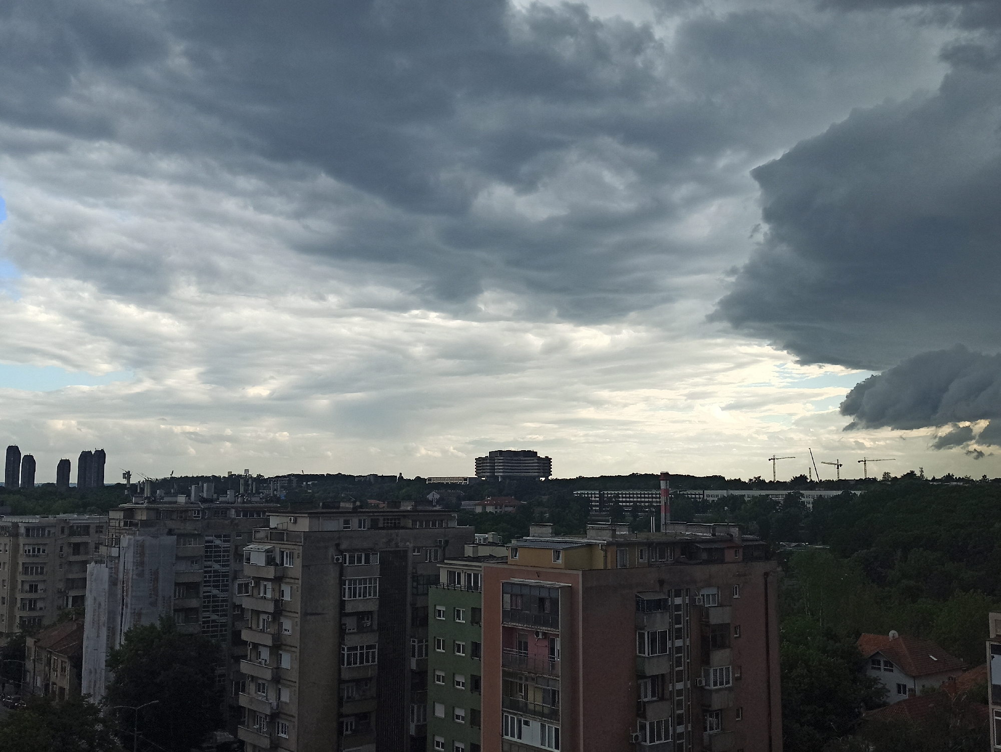 The image depicts a view of Vozdovac Urban Municipality, Serbia, under an overcast sky with storm clouds. In the foreground, a series of apartment buildings of varying heights and colors dominate the scene. Some are grey and appear older, while others are green, brown, or have other color accents. The buildings show signs of age and some repairs or construction. A few trees are scattered around the buildings. In the mid-ground, a row of trees separates the apartment complexes from the further landscape. Several taller buildings or structures are visible in the distance, along with construction cranes. The background shows a cityscape fading into the horizon.