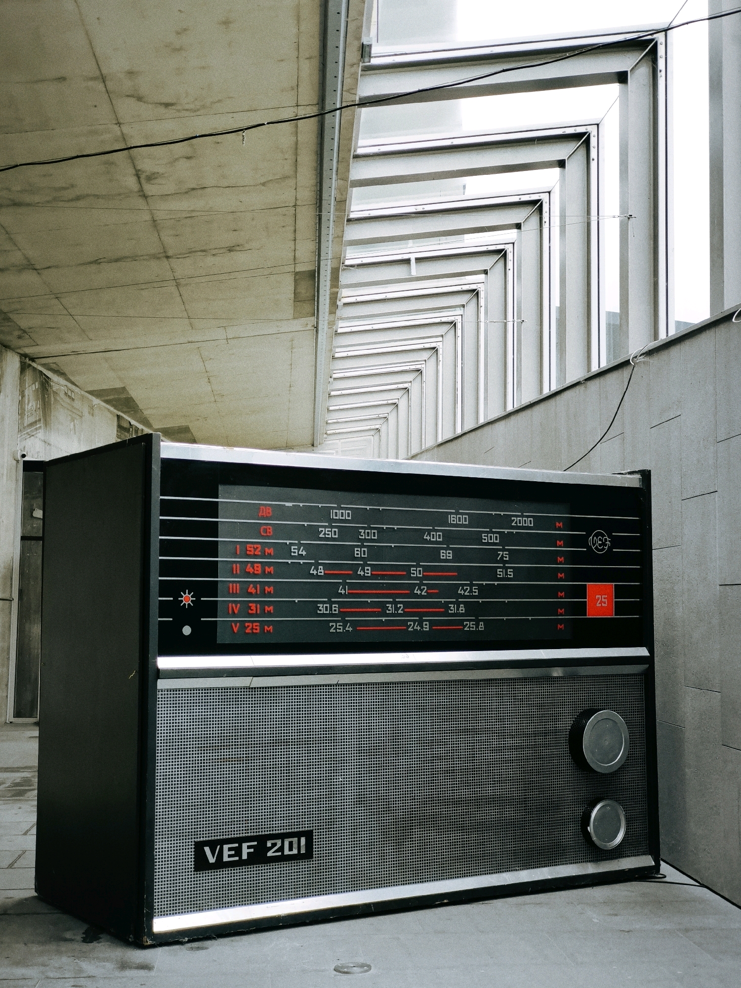 A large, vintage, black radio with a silver speaker grille and two silver knobs sits on a grey concrete floor. The radio has a display with red lettering and numbers. The background consists of modern architecture with concrete and metal elements, possibly an underpass or a covered walkway. The overall impression is one of an urban environment, perhaps a public space in the city of Tallinn, Estonia, given the style of architecture and the presence of an older piece of technology.