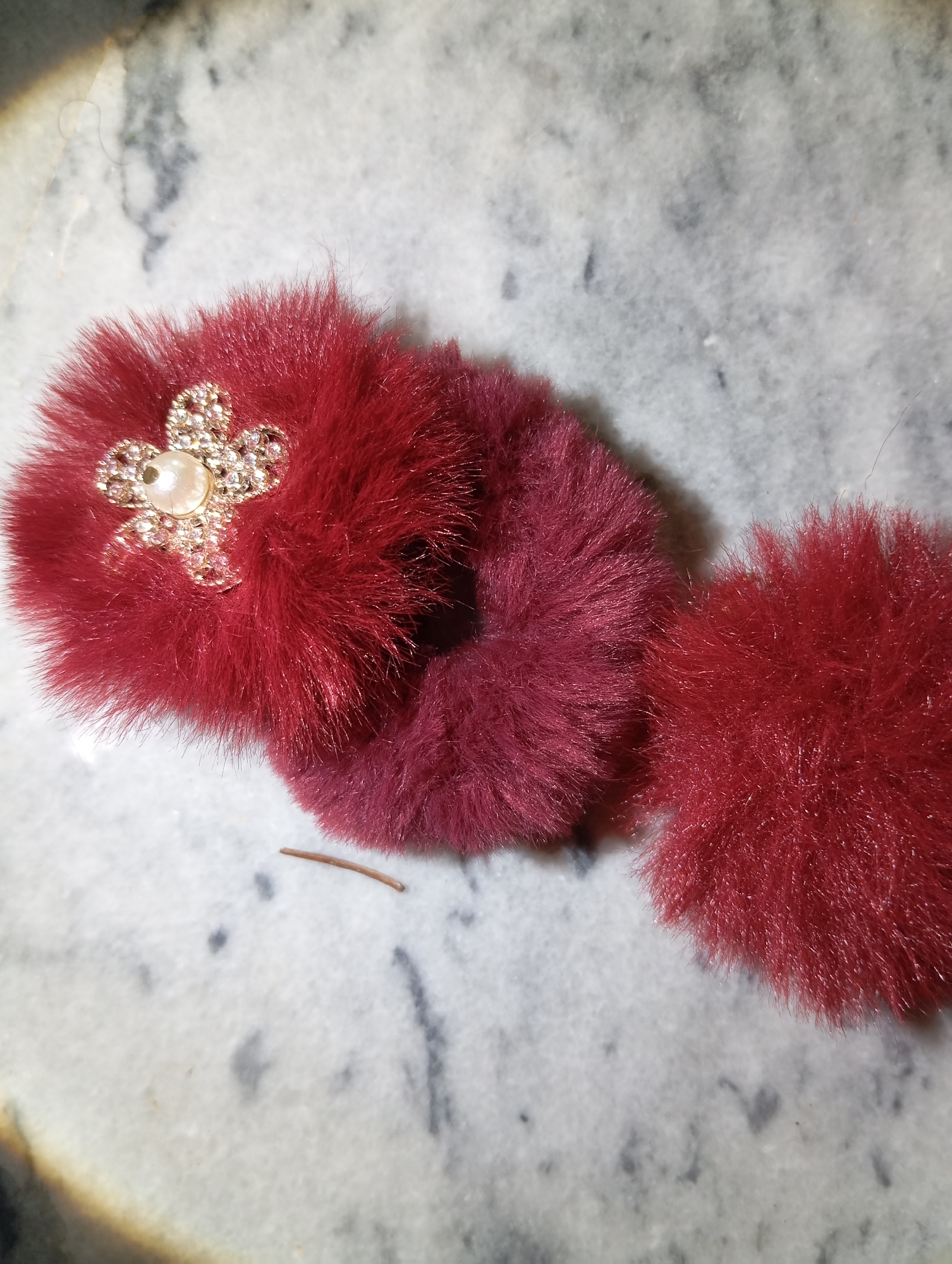 The image shows two plush, burgundy-colored hair accessories on a marble surface. The accessory on the left is a scrunchie decorated with a gold-colored floral charm, embellished with small clear rhinestones and a single pearl in the center. The accessory on the right is a fluffy pom-pom. A thin, brown twig lies on the marble surface between the two accessories. The background is a grey and white speckled marble pattern.