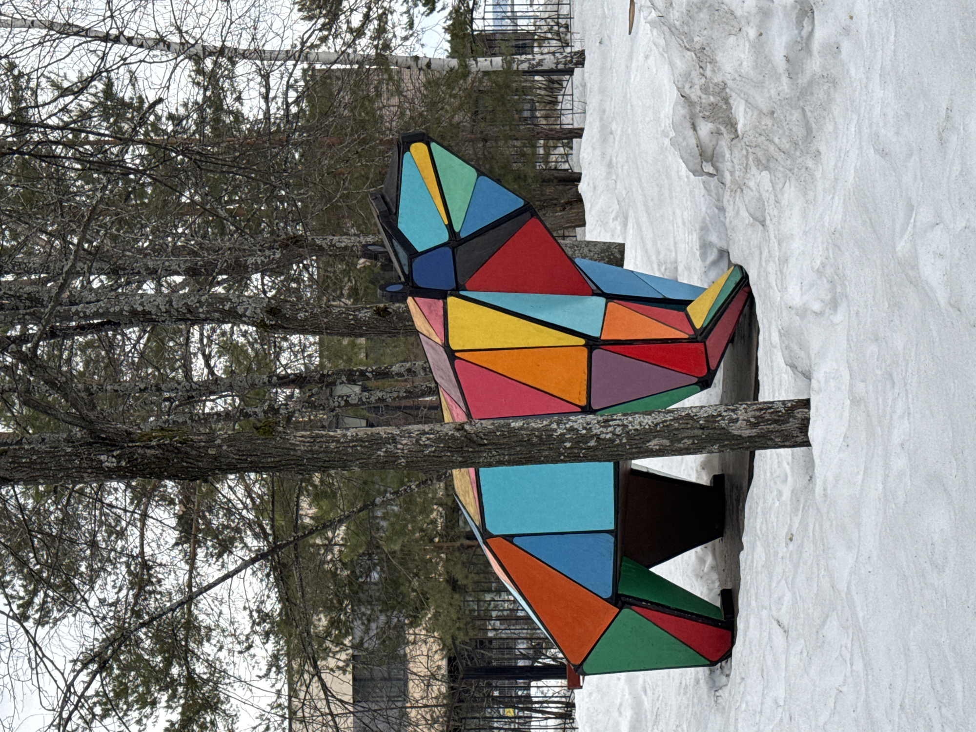 A colorful geometric sculpture is nestled between trees and snow. The sculpture is composed of many triangular and trapezoidal shapes, painted in a variety of bright colors including red, orange, yellow, green, and blue. The sculpture appears to be a public art installation, possibly located in the city of Chaikovsky, Russia. Bare tree branches are visible behind and around the sculpture. A snowdrift surrounds one side of the artwork. The sculpture sits close to the ground, and a tree trunk partially obscures the upper section.