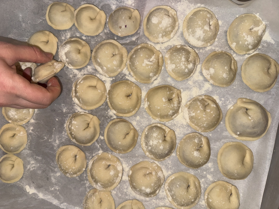 A person's hand holds a pelmeni, a filled dumpling, amidst a tray of many more. The dumplings are round, doughy, and feature a pinched top. Lightly dusted with flour, they are arranged on a sheet of parchment paper. The tray holds a variety of sizes and shades of the dumplings. The overall lighting is soft, accentuating the texture of the uncooked dough.