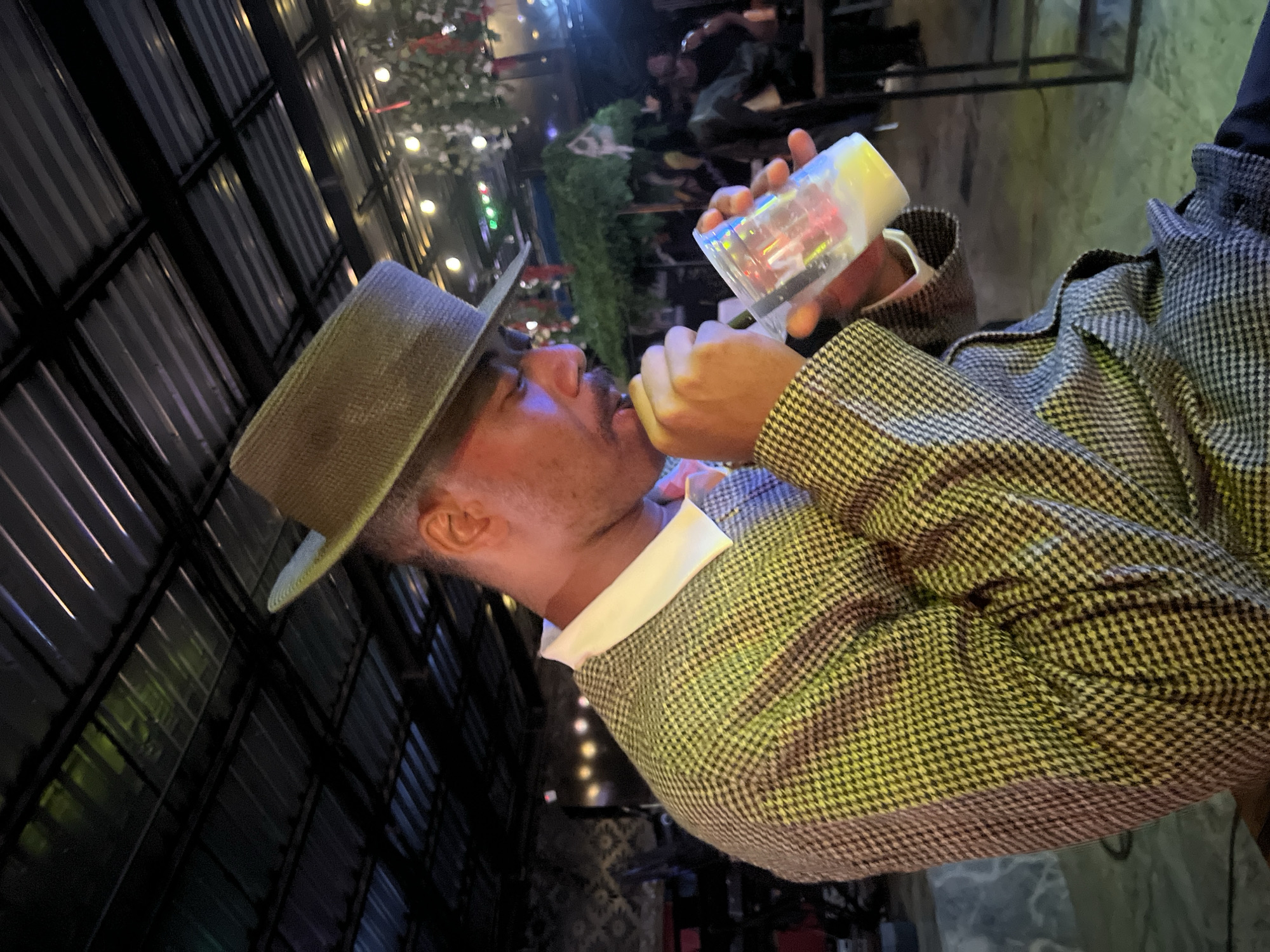 The image shows a man wearing a hat and a plaid jacket, drinking from a transparent cup. He is in an indoor venue with a corrugated metal roof and string lights. The man appears to be in a relaxed setting, possibly a bar or restaurant. The environment suggests a casual social gathering. The lighting is dim and atmospheric, with some green and yellow lights reflecting off the surfaces. The man is the central focus of the image, with his attention directed towards his drink. The background is slightly blurred, indicating the focus is on the man and his action. The overall impression is one of a person enjoying a moment of leisure in a dimly lit establishment.