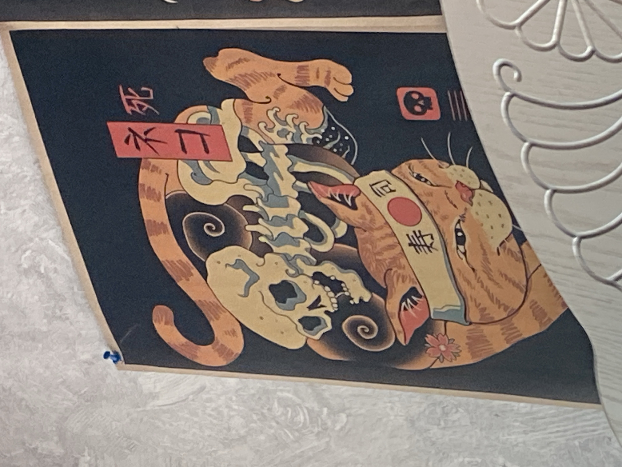 The image shows a section of a poster with Japanese-inspired artwork depicting a cat, a skull, and a snake. The poster is tacked to a textured wall with a blue thumbtack in the upper left corner. The artwork features an orange cat with a band around its head that has Japanese characters and a red circle, possibly representing a Hinomaru. A stylized skull is shown behind the cat, intertwined with what appears to be a snake or a swirling pattern. The background of the poster is dark blue. There are also small icons in the lower right section of the poster, including a skull within a red square and some horizontal lines. A white decorative metal railing is visible in the foreground, partially obscuring the bottom of the poster. The location is identified as Shakhtinsk, Kazakhstan, but there are no visual cues in the image to confirm this specific location.