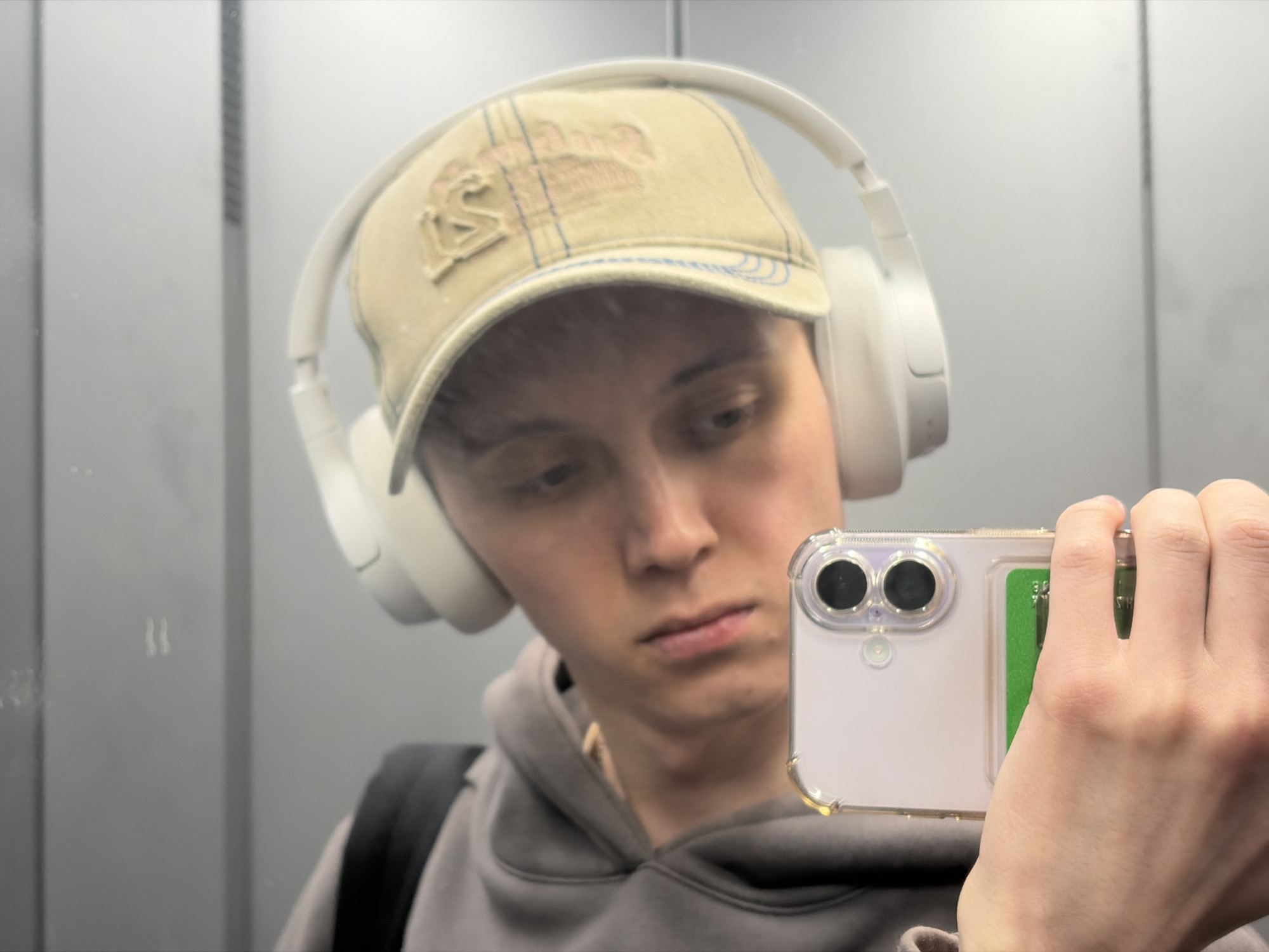 A young person with short brown hair, wearing a tan baseball cap with an embroidered logo and white over-ear headphones, is taking a selfie in an elevator. They are looking down at the phone in their right hand, which has a clear case with a green card in the back. They are also wearing a gray hoodie and a black backpack. The elevator walls are a reflective gray, with vertical seams visible.