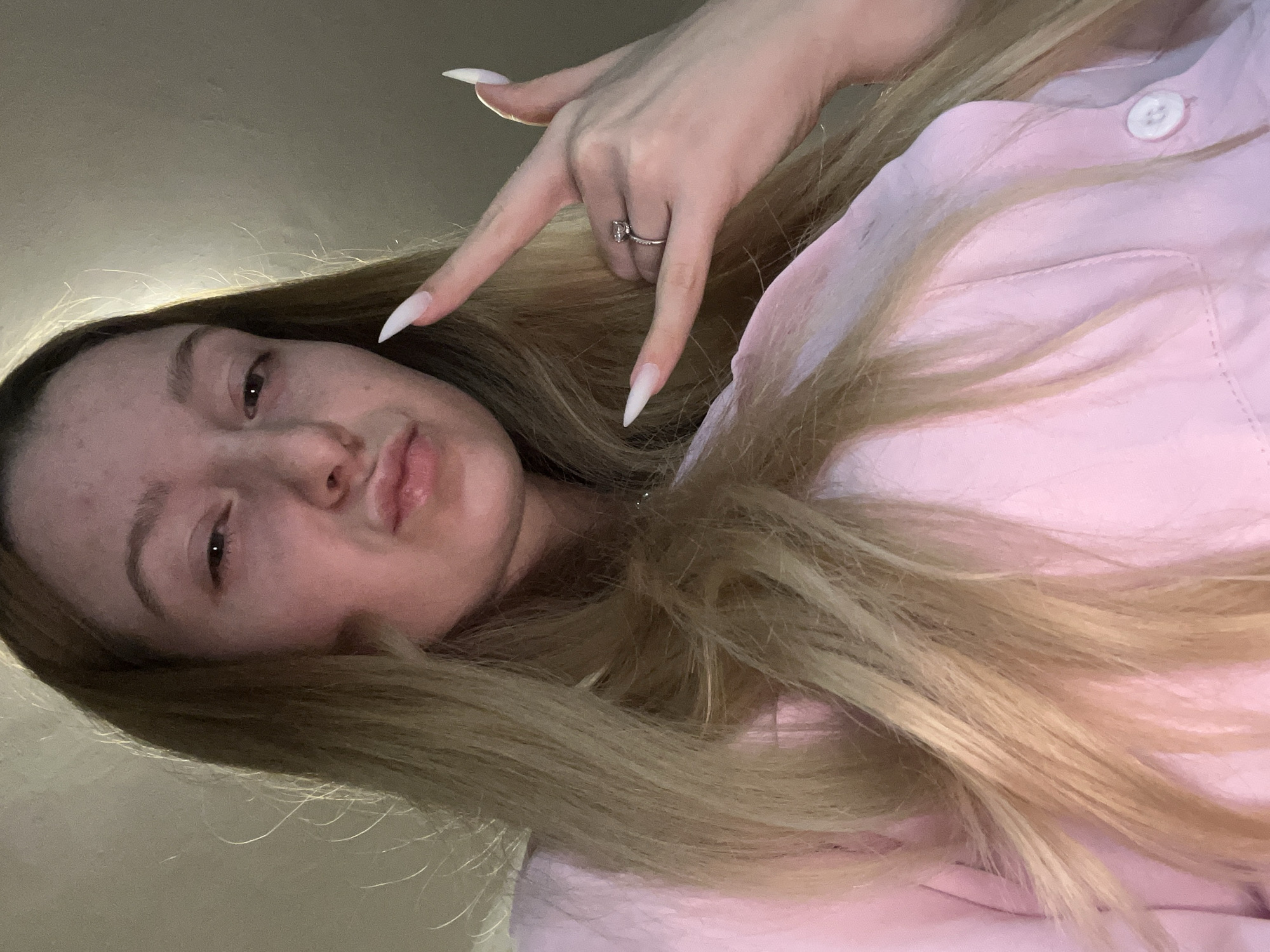 A young woman with long blonde hair, wearing a pink shirt, poses for a selfie. She is holding up her hand in a rock-on gesture, and a ring is visible on her finger. The background is a plain, light-colored wall. The image appears to be taken indoors. The location in the city of Varna, Bulgaria is not discernible.