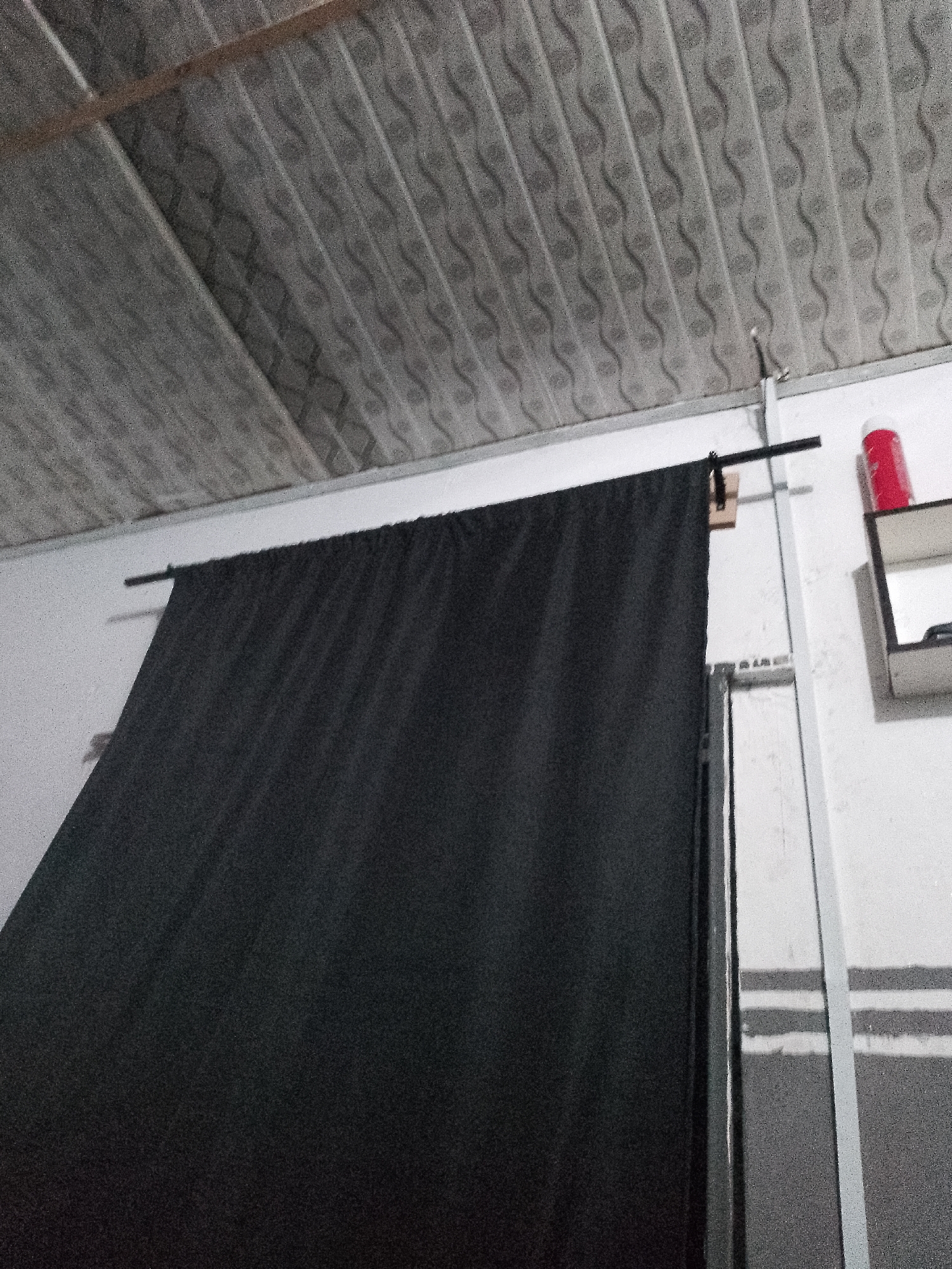 The image shows a close-up of a dark curtain hanging from a rod that is attached to a white wall. The ceiling above is paneled with a repeating wave pattern. To the right of the curtain, a red cylindrical object is visible on a shelf. The scene appears to be indoors, possibly a studio or a room with a makeshift backdrop. There are no people or any indication of a specific location within a city.