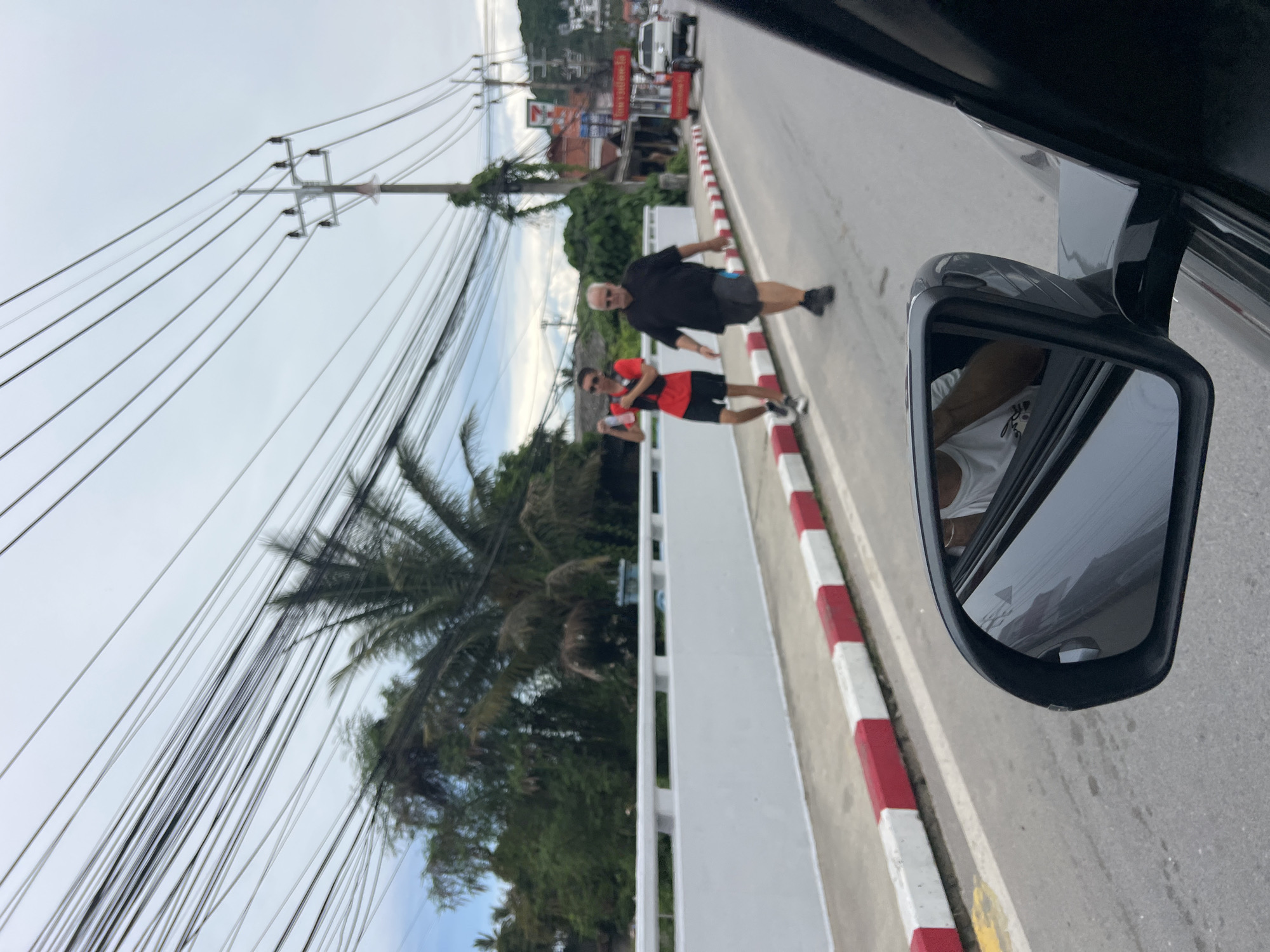 In Baan Hua Thanon, Thailand, a man in a black t-shirt and shorts walks on the right side of the road, while another man in a red t-shirt and black shorts walks behind him on the left side of the road. Both men are walking away from the viewer. The man in red is holding a water bottle. They are walking on a paved road with a white and red striped curb on the left. In the background, palm trees and buildings line the street, with power lines overhead. A white car and a red cart are also visible in the distance. The scene appears to be captured from a vehicle, as a car's side mirror is visible in the foreground. The sky is overcast.
