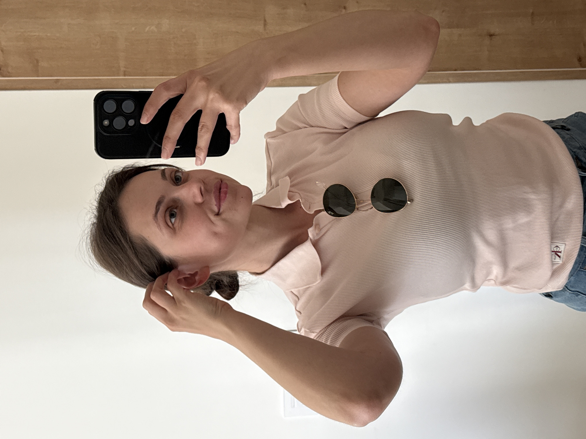 A woman is taking a selfie in a mirror. She is holding a black smartphone in her right hand, with the camera directed towards her face. She is wearing a light pink ribbed polo shirt and sunglasses are positioned on the shirt near her right shoulder. Her left hand is near her ear. The background consists of a white wall and a wooden surface above it. The location is not evident from the image and cannot be determined to be Belgrade, Serbia.