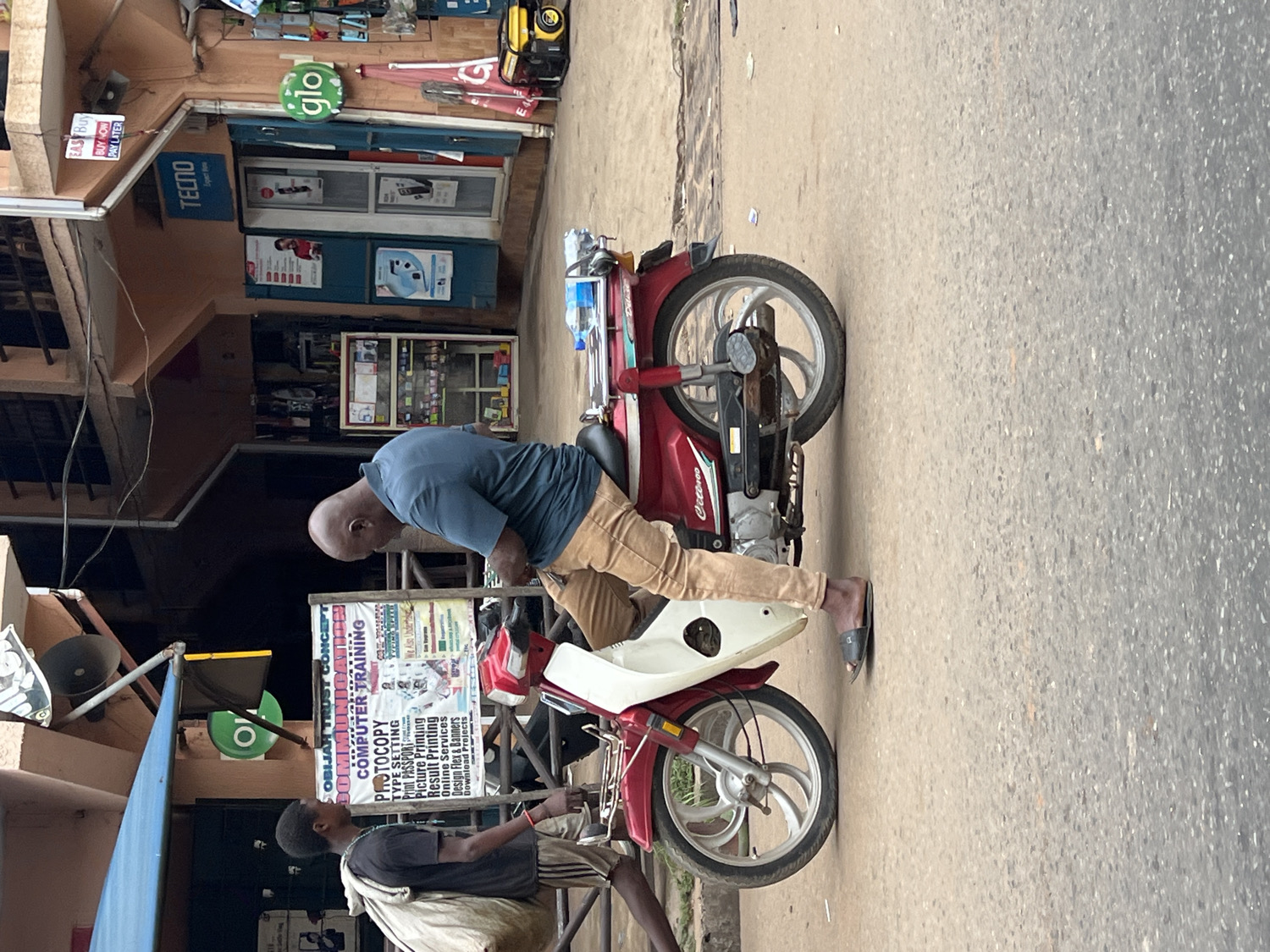 This image captures a daytime street scene in Nkpor, Nigeria, featuring two males and a motorcycle parked on a paved road next to commercial buildings.
In the mid-ground, a bald man wearing a blue t-shirt and tan pants is straddling a red and white motorcycle. He is leaning forward, looking down, possibly at a younger boy who stands in front of him. The man is wearing black flip-flops on his feet, with one foot on the ground. The motorcycle has "Critereos" written on its side and a plastic water bottle placed on its rear.
To the left, a younger boy with a dark t-shirt and striped shorts is standing near the front wheel of the motorcycle, facing the older man. He appears to be holding a light-colored bag or cloth.
The background reveals several shopfronts. One prominent sign features the "TECNO" logo with "Expert Poe" below it, indicating a mobile phone or electronics store. Another shop displays a bright green "glo" logo, belonging to a telecommunications company. A sign above a shop reads "BUY NOW PAY LATER".
A large, detailed sign near the younger boy lists various services under "OBIJAH TRUST CONCEPT," including "COMPUTER TRAINING," "PHOTOCOPY," "TYPE SETTING," "Print PASSPORT," "Picture Printing," "Result Printing," "Online Services," "Design Flex & Banners," and "Download Projects." The overall impression is that of a bustling commercial area. The weather appears clear, with bright, natural lighting.