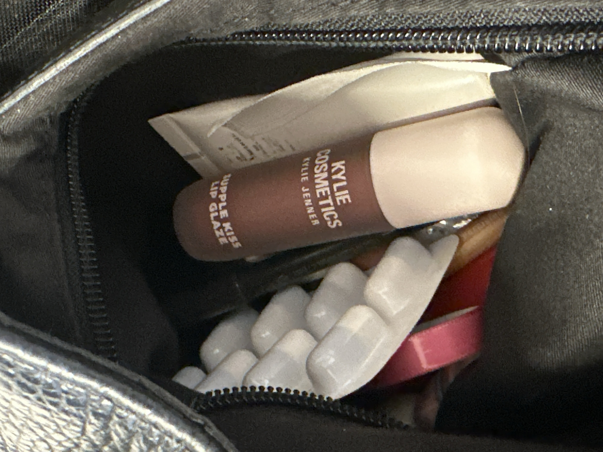 The scene depicts the interior of an open, dark-colored bag with a textured exterior, located in the city of London, United Kingdom. A black zipper runs along the bag's opening. Visible contents include a cylindrical Kylie Cosmetics product, specifically labeled "KYLIE JENNER KYLIE COSMETICS RUPPLE KISS LIP GLAZE" (text inverted), which features a dark brown body and a light beige base or cap. To the left of this product, a white, translucent strip is visible, containing a series of interconnected, elongated objects, resembling false nail tips or a blister pack. Partially obscured behind the Kylie Cosmetics item, a white pouch or tube is present. A narrow, dark pink ribbon or strap is also partially visible beneath the white translucent strip. No individuals are present in the frame, and no actions are depicted beyond the static arrangement of these items within the bag.