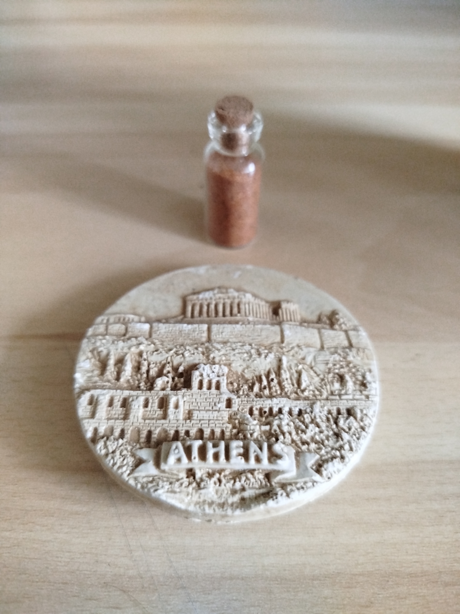 The image shows a small glass bottle with a cork stopper, filled with a reddish-brown substance. In front of the bottle is a circular, off-white souvenir depicting the city of Athens, Greece. The souvenir has raised details showing buildings and the word "ATHENS". The objects are placed on a light brown, possibly wooden, surface. The image does not depict anything relating to the city of Varna, Bulgaria.