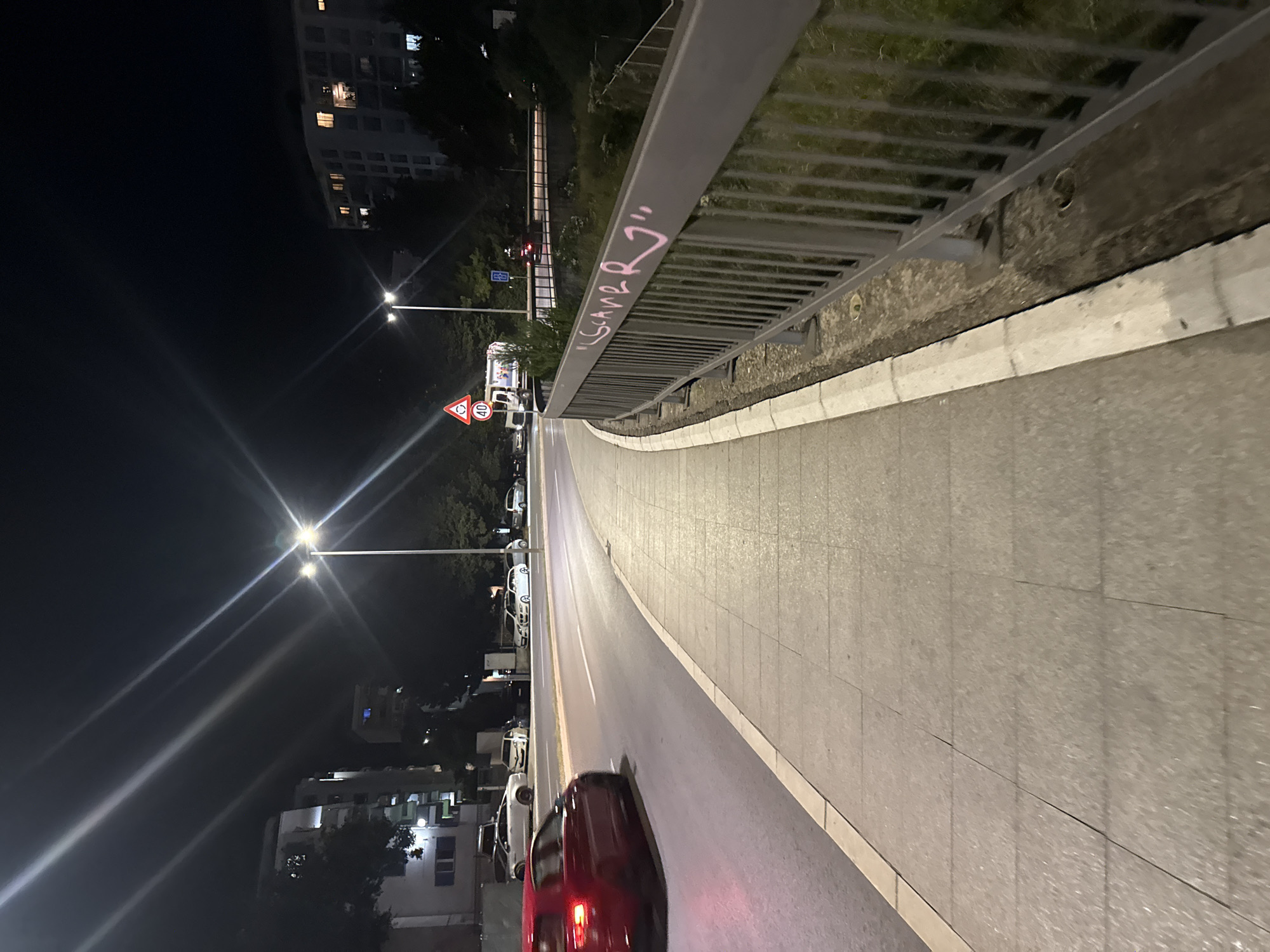 The image depicts a nighttime street scene in Boreti, Montenegro. A red car is captured in motion on the left side of the frame, driving on a paved road. To the right of the road is a sidewalk made of grey rectangular tiles. A metal railing runs along the edge of the sidewalk, and pink graffiti is visible on it, reading "LCSCAR RJ". Streetlights illuminate the scene, casting long streaks of light across the dark sky and onto the road and sidewalk. In the background, buildings and trees are visible, along with other vehicles and street signs, including a speed limit sign indicating 40. The overall environment appears to be a well-lit urban area at night.