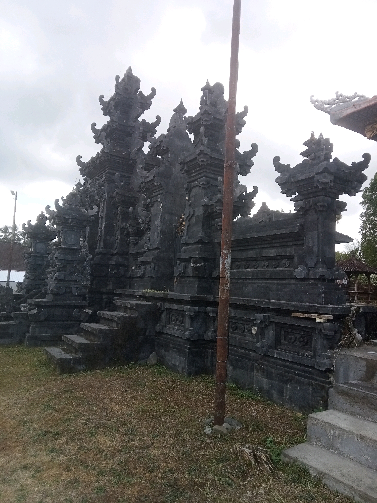 An overcast day in Mendoyo, Indonesia, features a stone temple entrance with intricate carvings. The structure is dark gray, with multiple tiered roofs and ornate decorative elements. Steps lead up to the entrance on the left side of the frame. A tall, rusty metal pole stands prominently in the foreground, slightly off-center. The ground is covered with dry grass and scattered weeds. In the background, a few trees and a distant building are visible.