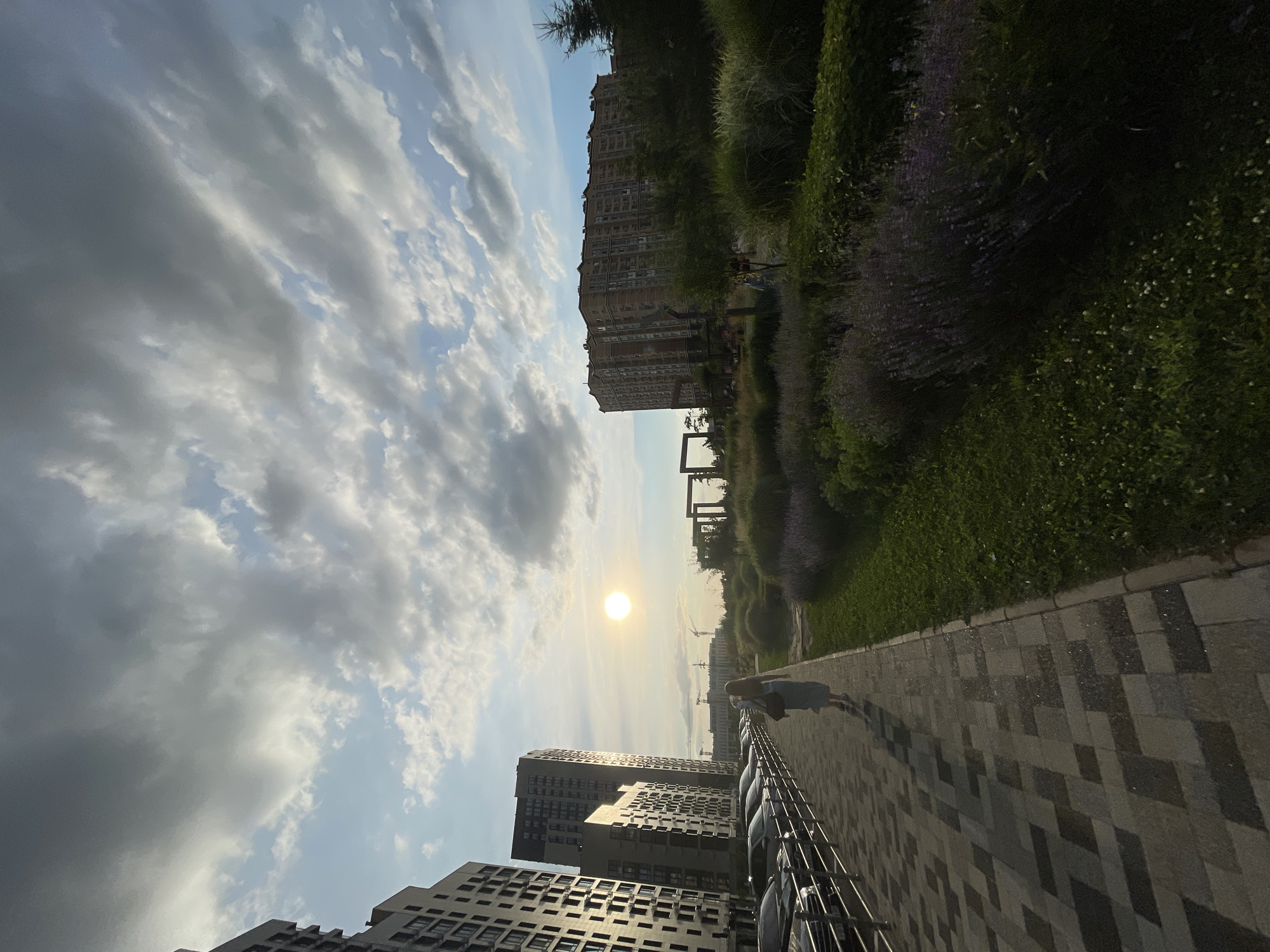 The image depicts an outdoor scene in an urban environment. A woman wearing a light blue dress and carrying a bag walks along a paved walkway. To her left, there is a row of parked cars bordered by a metal fence. Beyond the cars, several multi-story apartment buildings with numerous windows are visible. To the right of the walkway, there is a grassy area with various plants, including lavender. In the background, a large, light-colored apartment building stands, and further in the distance, construction cranes can be seen. The sun is visible in the sky, partially obscured by clouds, creating a bright glow. The sky is partly cloudy with a mix of gray and white clouds. The scene appears to be set in a residential area with landscaping and architectural elements. The location is identified as the city of Stavropol, Russia.