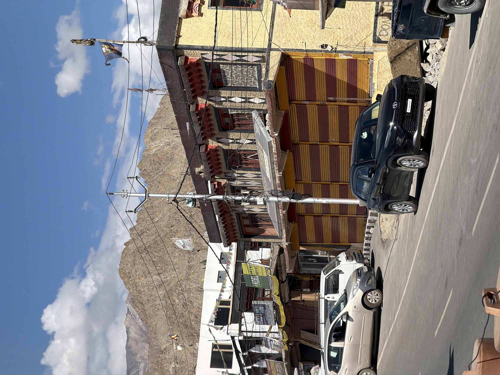 The image captures a street scene in Leh, India, on a bright, sunny day. The foreground features a paved road with white lane markings, and several vehicles are present. A light-colored hatchback is parked on the left side of the road, facing away from the viewer, with a white van behind it. A black SUV is positioned in the right lane, facing towards the viewer. The background shows a row of buildings with distinctive architecture, featuring yellow facades, red trim, and what appear to be roll-up shutters painted in alternating bands of red and yellow. Prayer flags, common in the region, are visible strung between buildings and on poles. Beyond the buildings, rugged, arid mountains rise under a clear blue sky with scattered white clouds. Power lines crisscross the scene, supported by utility poles. A few signs are affixed to the buildings, though most are difficult to read. The overall impression is of a typical street in Leh, characterized by its unique blend of traditional architecture, mountainous terrain, and everyday urban activity.