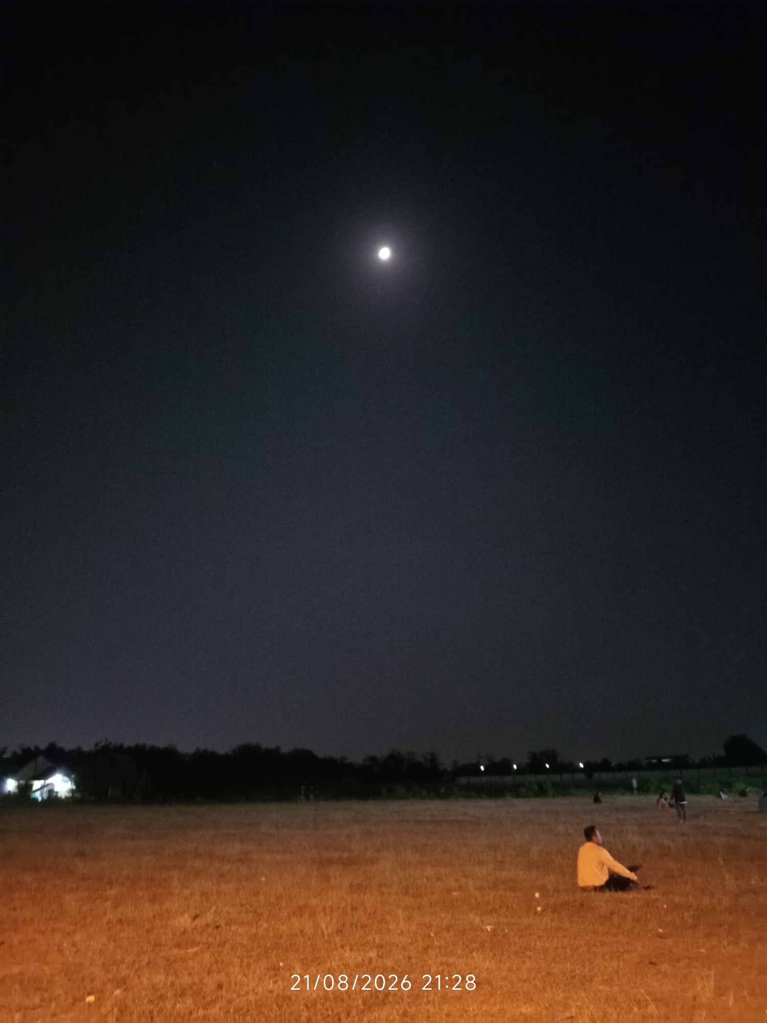 The image depicts a wide, flat, dry grassy field at night. A bright full moon is visible in the dark sky. In the foreground, a single person is sitting in the grass, facing away from the camera, wearing a light-colored long-sleeved shirt. In the background, a line of trees forms a dark silhouette against the horizon, with some lights from buildings visible behind them. A date and time stamp, "21/08/2026 21:28", is present in the lower right corner of the image. The overall scene suggests an outdoor gathering or leisure activity in a semi-rural or suburban area near Canggu, Indonesia.