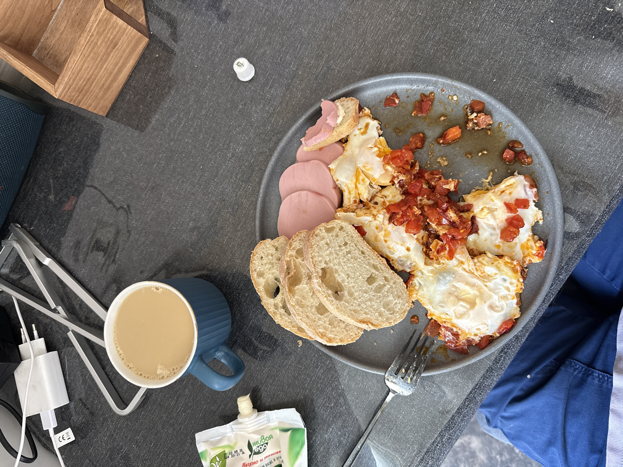 The image displays a meal set on a dark grey, textured surface, likely a table, in Khimki, Russia. A person is partially visible in the lower left, wearing blue trousers and having a grey fabric on their lap. A round, dark grey plate centrally holds fried eggs mixed with diced red vegetables (likely tomatoes or peppers), three slices of light-colored bread, and several slices of round, pale pink processed meat. A small piece of bread with a pinkish spread is also on the plate. A metal fork rests on the left side of the plate. In the upper left, a ribbed, teal-blue ceramic mug contains a light brown, frothy beverage. Adjacent to the mug, a white and green squeeze pouch with Cyrillic text, reading "МЯСНОЙ РЯД" and "ВКУСНО и ПОЛЕЗНО," dispenses a small dollop of white substance. The background includes white charging cables connected to a white power adapter, a silver-colored metal stand, a partial view of a teal speaker with a textured grill, and a light brown wooden multi-compartment box. A small white plastic cap is visible on the table surface in the mid-right. The scene indicates a breakfast or meal in progress.
