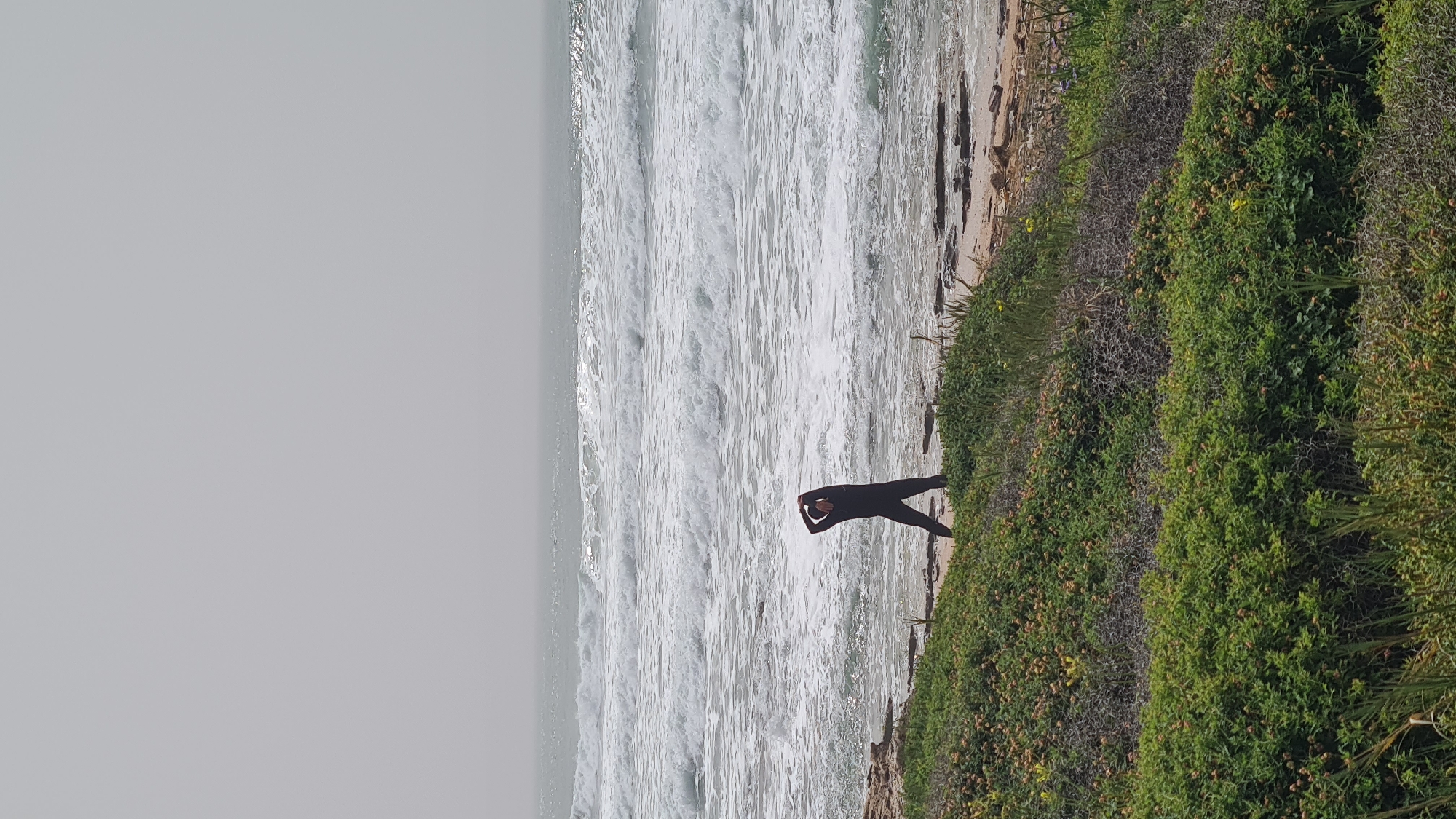 A medium shot depicts a person in a black wetsuit stretching on a verdant cliffside overlooking a beach and ocean. The sky is overcast, lending a muted tone to the scene. The beach appears sandy with visible waves. The vegetation on the cliffside is dense and primarily green. The subject's pose suggests preparation for activity or relaxation. It is daytime. The location is Nahariyya, Israel.